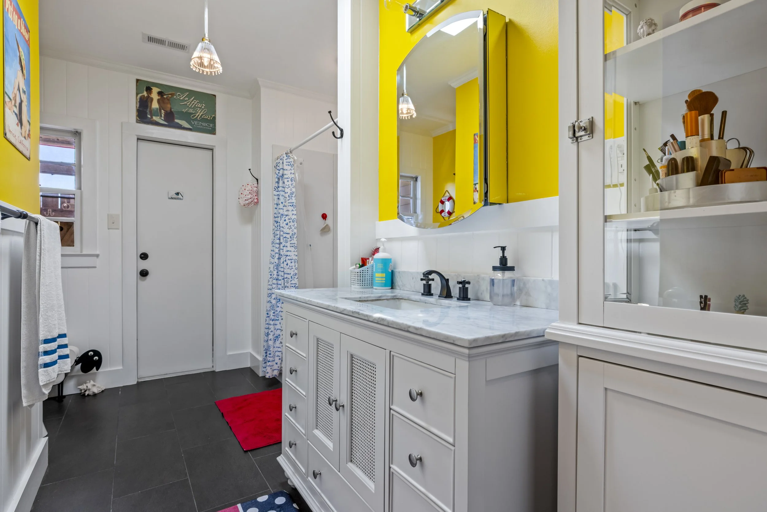 A bathroom with white cabinetry, a marble countertop, yellow accent wall behind a mirror, a white medicine cabinet, and a countertop with various toiletries. There is a shower with a blue and white patterned curtain, a white door with a small window,