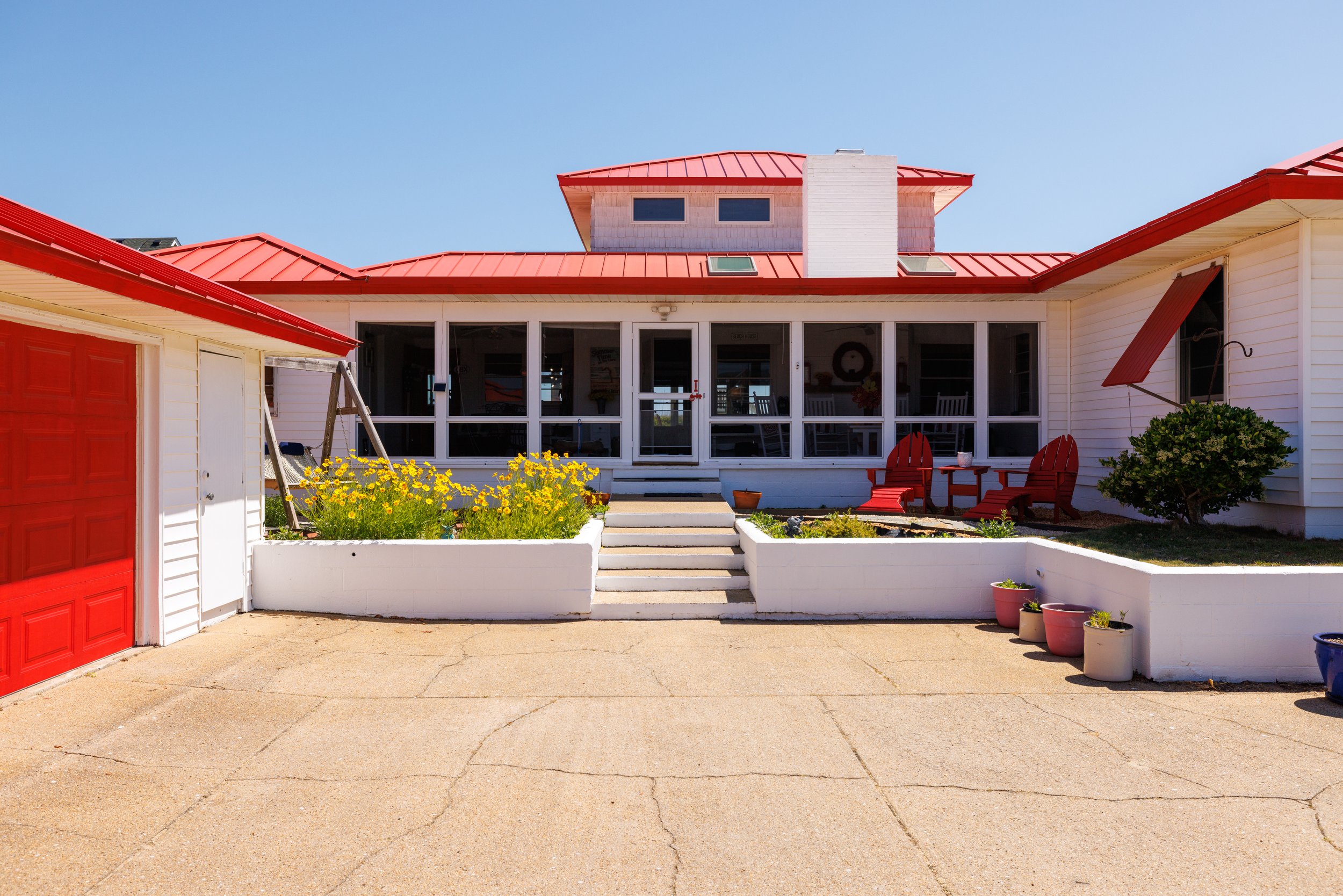 View of a house with a red metal roof, large screened porch with two red Adirondack chairs, and potted plants. The scene includes a concrete driveway and a clear blue sky.