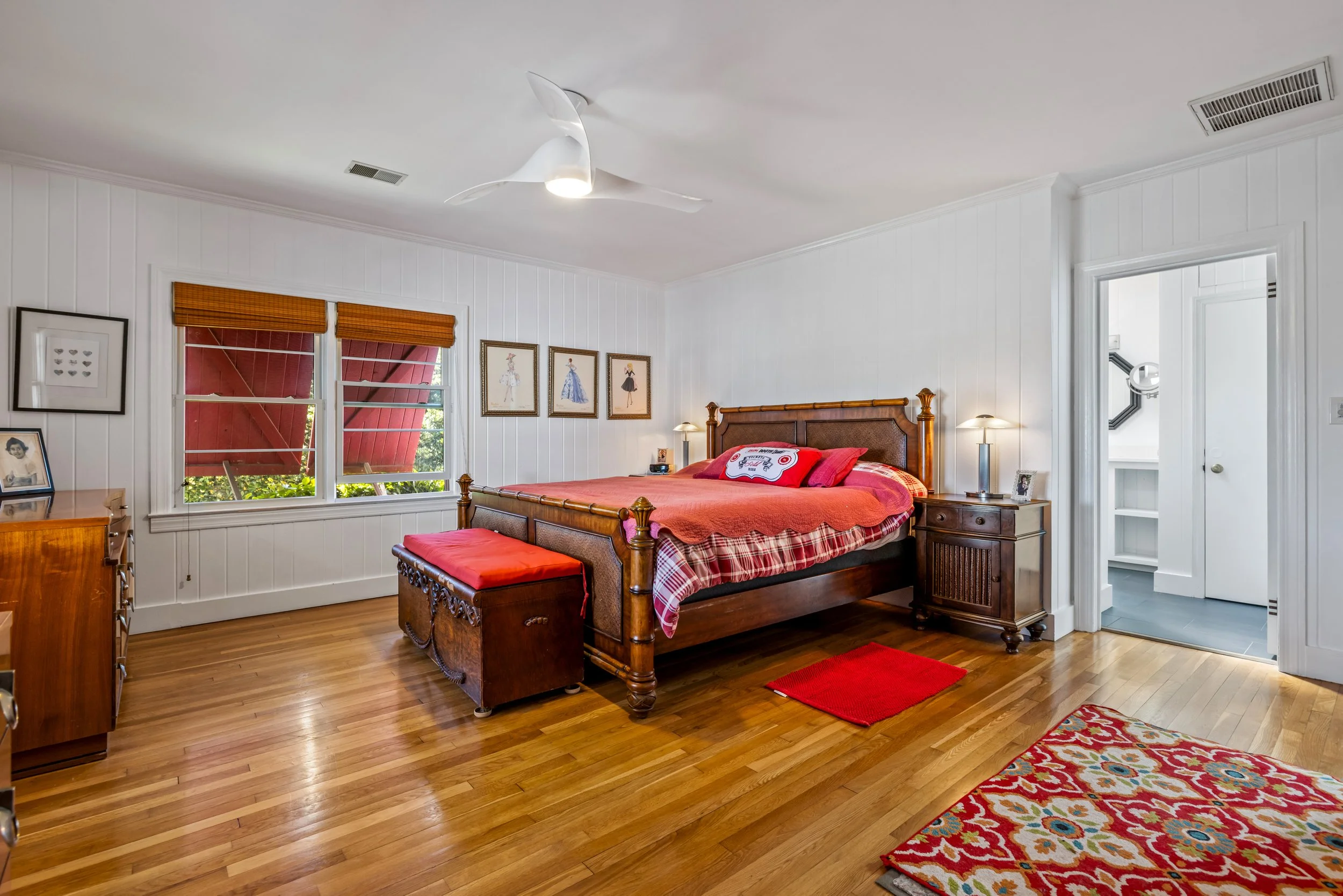 Bedroom with wooden floor, decorated with framed pictures, a bed with reddish-orange bedding, two bedside tables with lamps, a colorful rug, and two windows with wooden blinds.