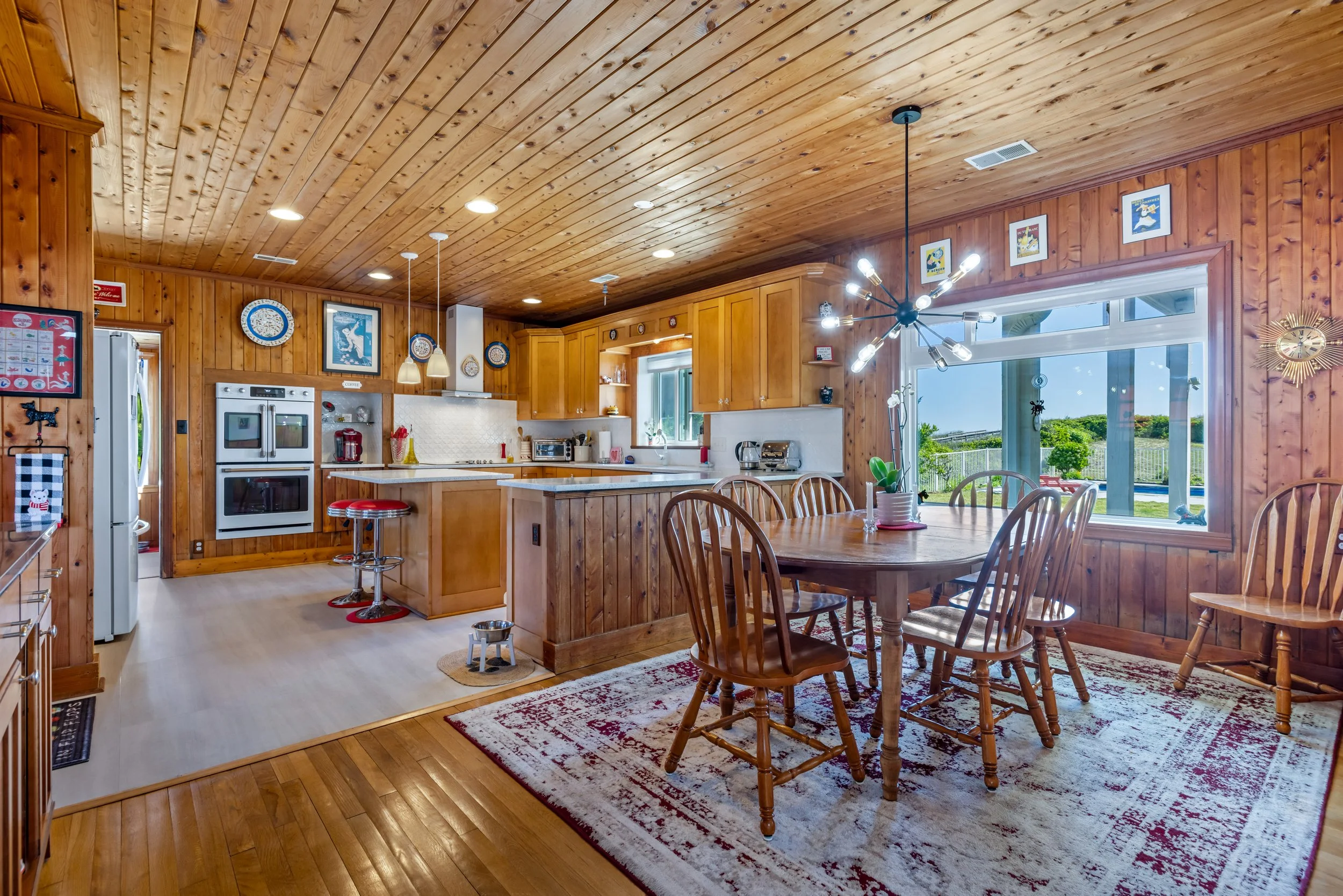 A room with wooden walls and ceiling, a dining table, chairs, kitchen appliances, and large windows showing an outdoor view.