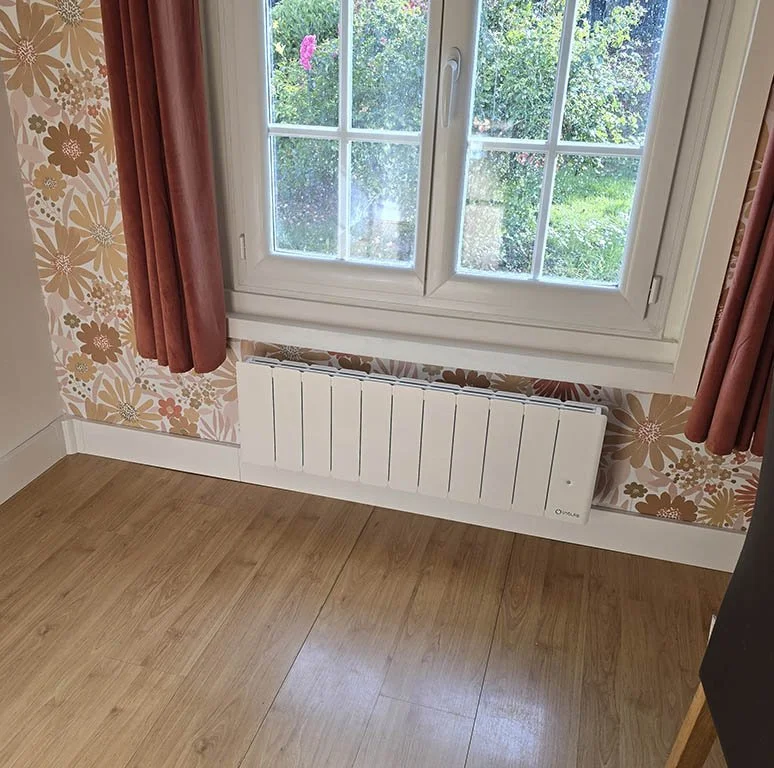 Un radiateur connecté Intuis Beladoo Nativ modèle plinthe installé sous une fenêtre dans un chambre