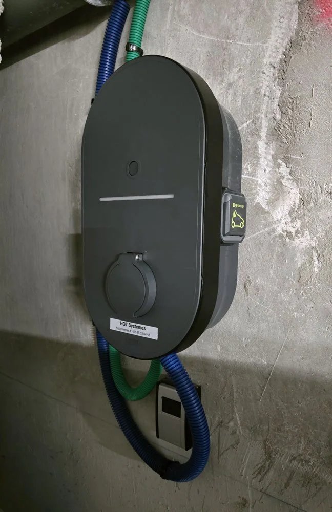 Un panneau de contrôle pour une station de recharge de véhicule électrique, avec un bouton de contact, des câbles bleus et verts, fixé au mur en béton.