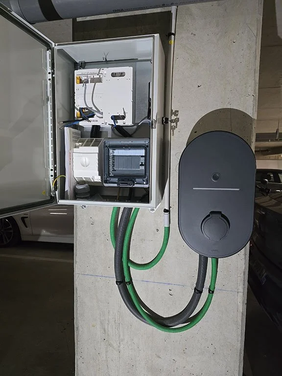 Une armoire électrique ouverte contenant des composants électroniques, connectée à un boîtier noir avec des câbles vert et noir qui sortent, située dans un parking souterrain.