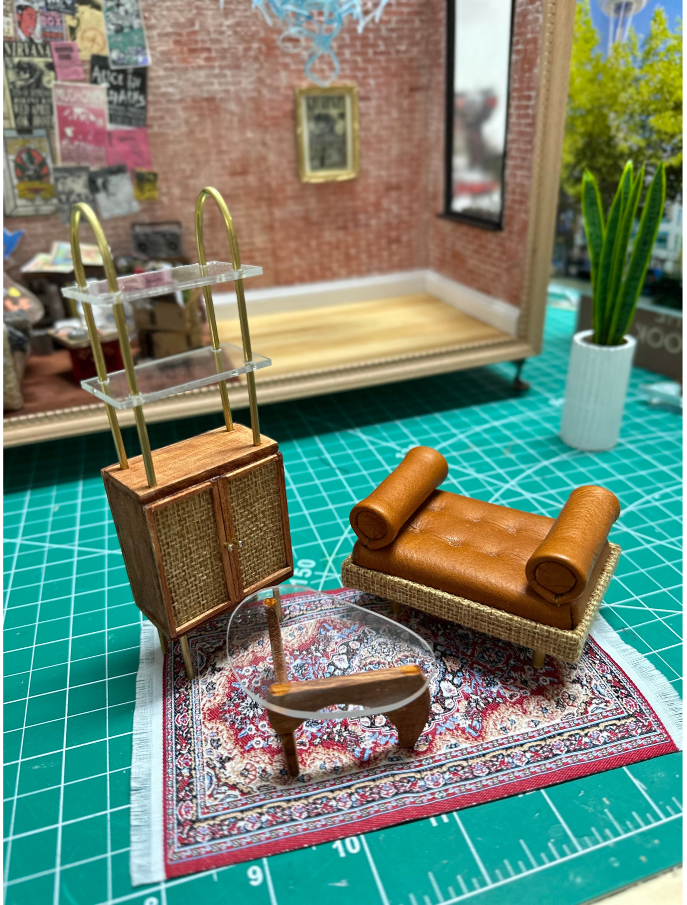 Miniature living room scene with a tiny sofa, a small round table, a woven cabinet with gold handles, and a tall houseplant, set on a patterned rug against a brick wall with posters and a framed picture.