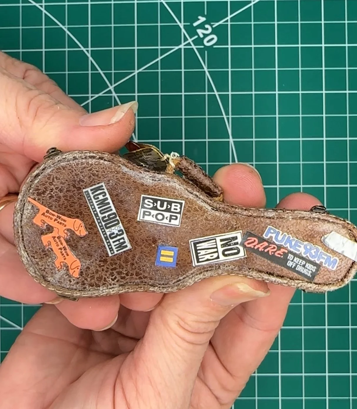 A person's hand holding a brown shoe with various brand and logo stickers on the sole, with a green cutting mat with grid lines in the background.