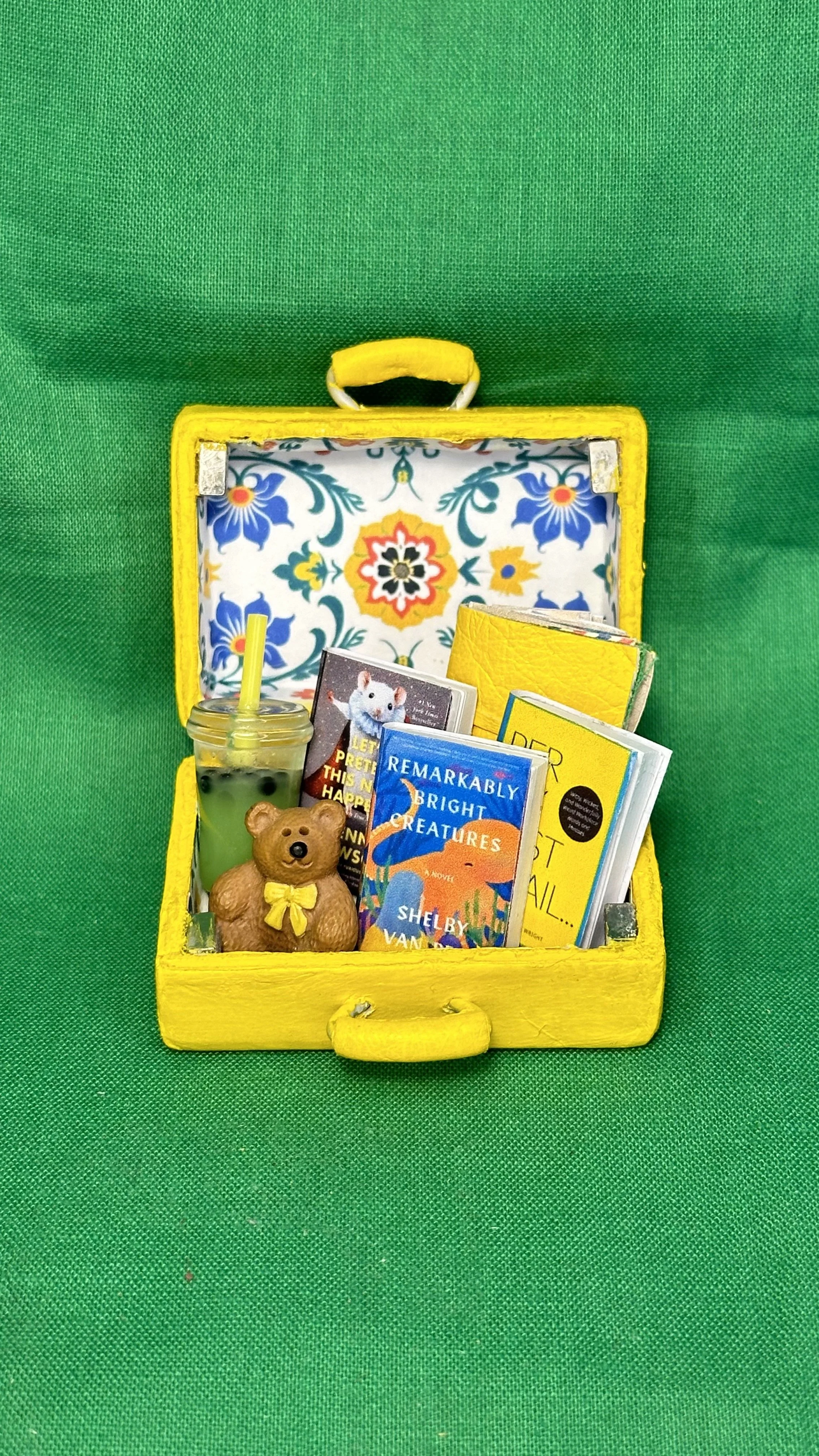 A small, open yellow toy suitcase with a floral interior lining, containing children's books, a teddy bear, a water bottle with a yellow straw, and a small figurine of a bear with a yellow bow, set against a green background.
