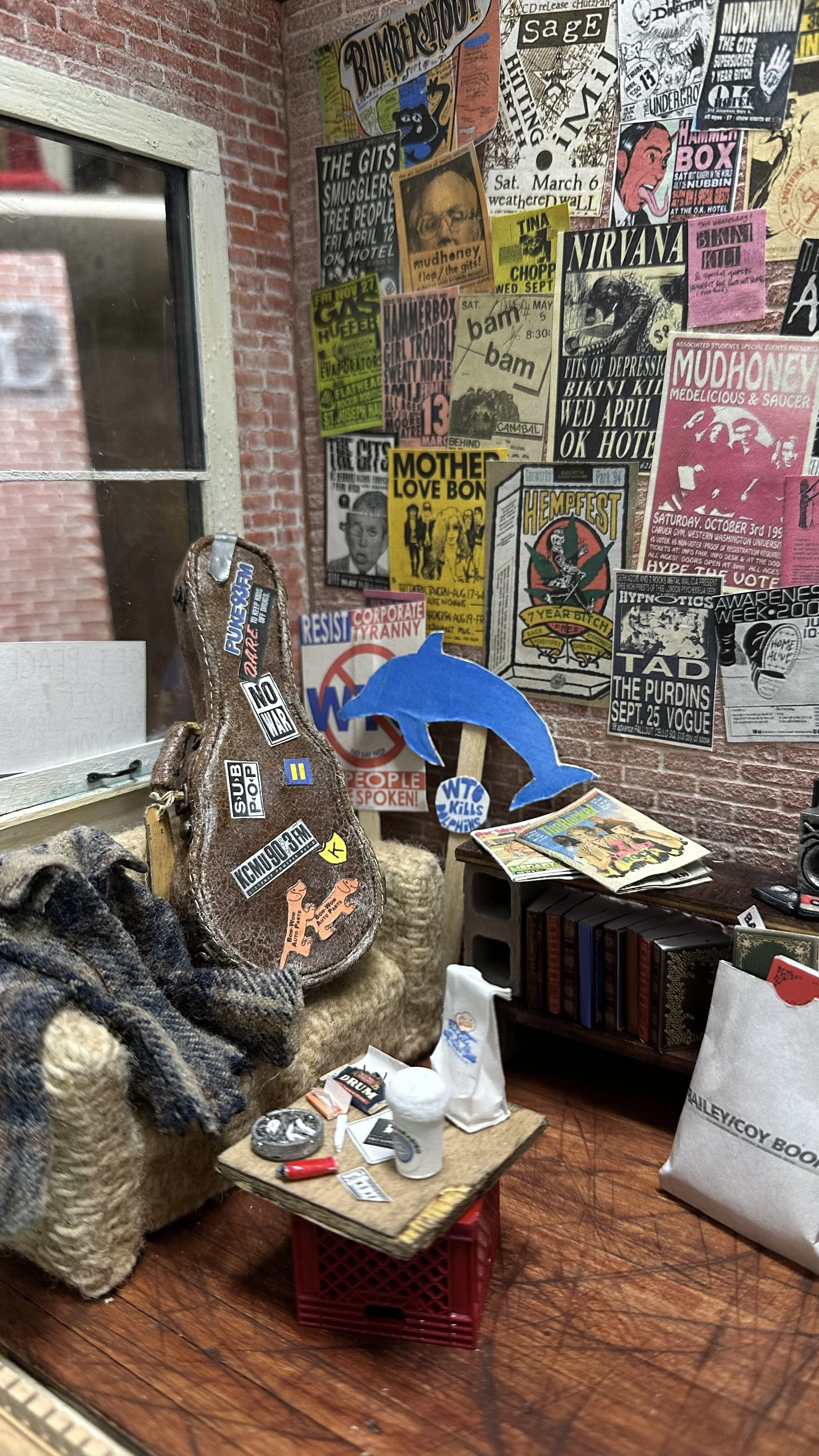 A small table with various items including a white cup, a small round mirror, and a lighter, placed on a red plastic crate. In the background, a collage of protest posters and flyers covers a brick wall. To the left, part of a window with a white fra