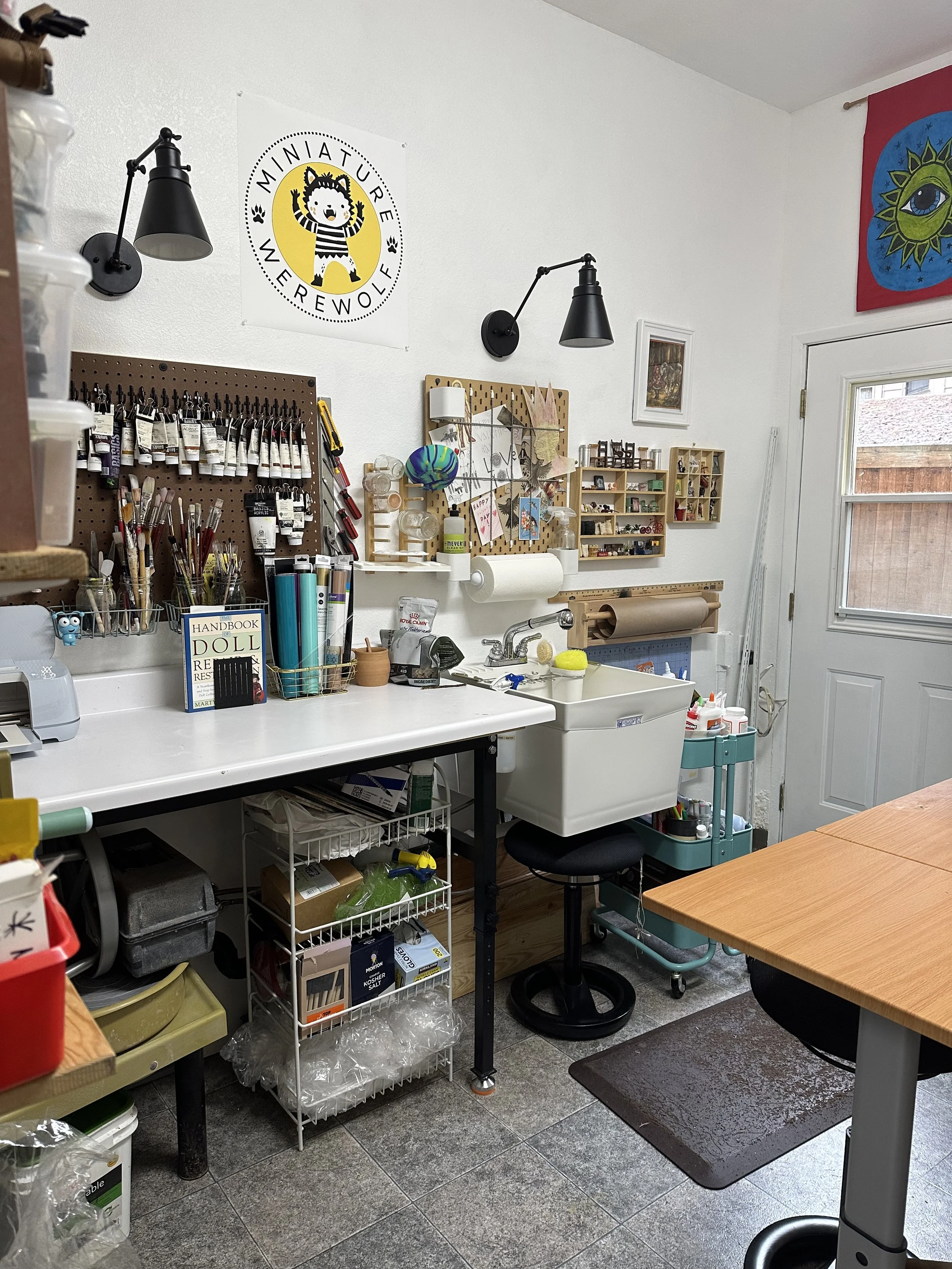 A craft room featuring a wall with art supplies, including paint tubes, brushes, and a pegboard with notes and decorations. There are shelves with various small items, a sink under a window, and a table with supplies. The room has a white wall with a Minature Werewolf logo and a colorful tapestry with a sun and eye design.
