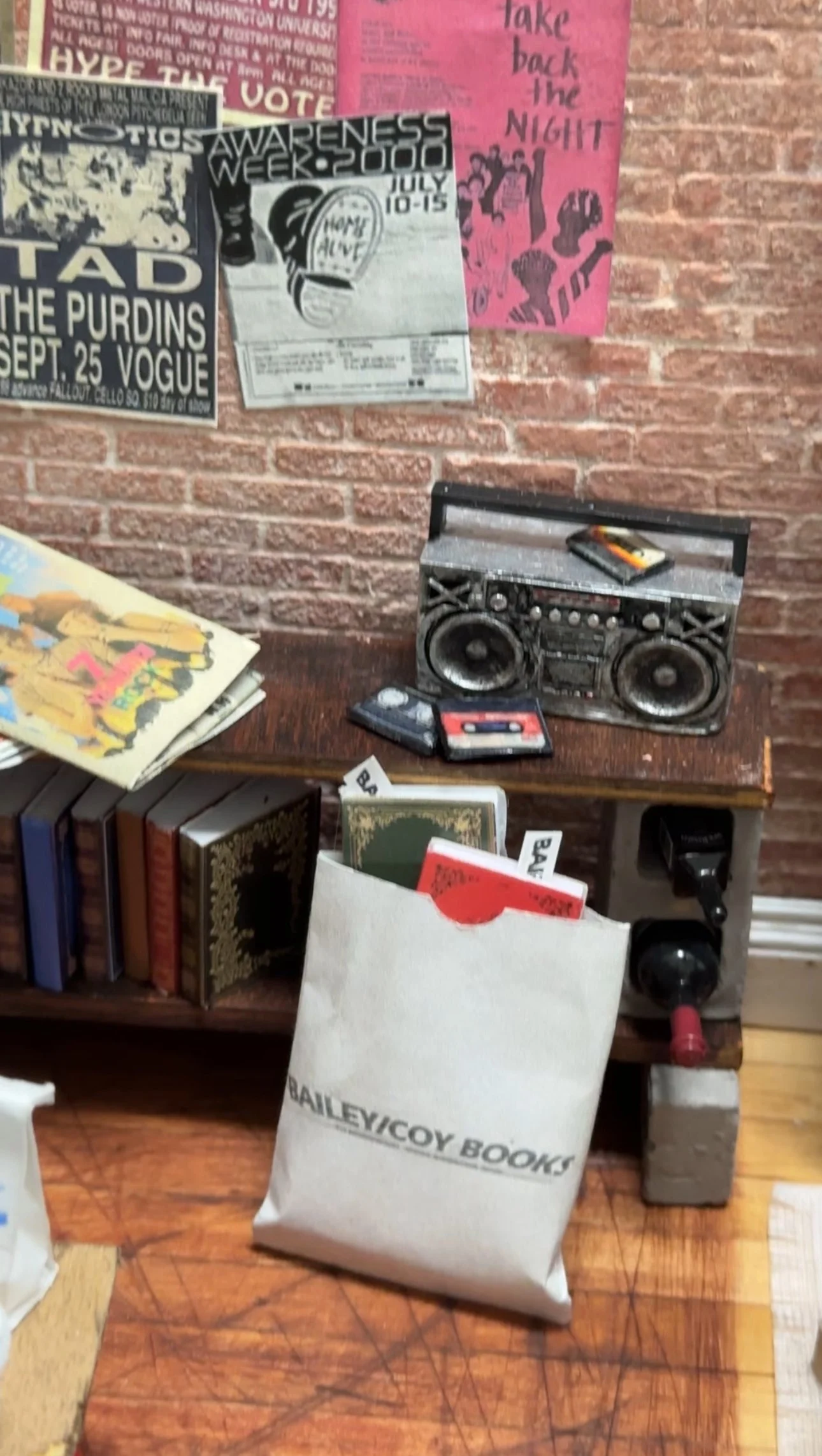 A cluttered wooden table with books, a white paper bag, and a small boombox against a brick wall decorated with posters.