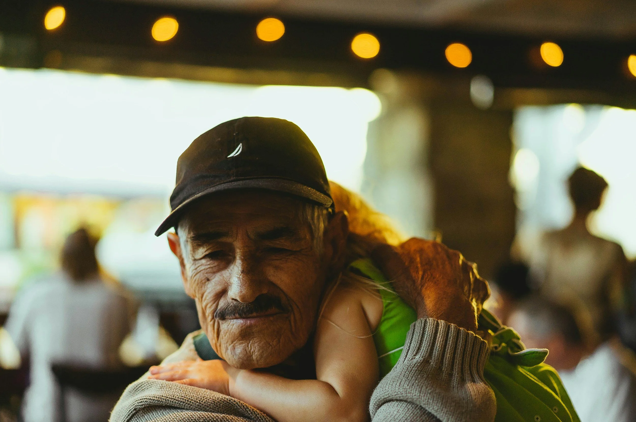 An elderly man with a dark gray baseball cap is hugging a young person who is wearing a green shirt. The scene appears to be in a lively outdoor setting with string lights overhead and other people blurred in the background.