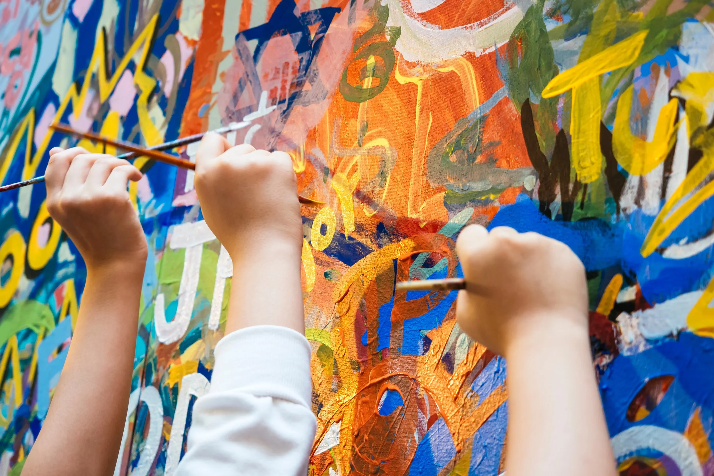 Close-up of several children's hands painting on a colorful mural with abstract shapes and bright colors.