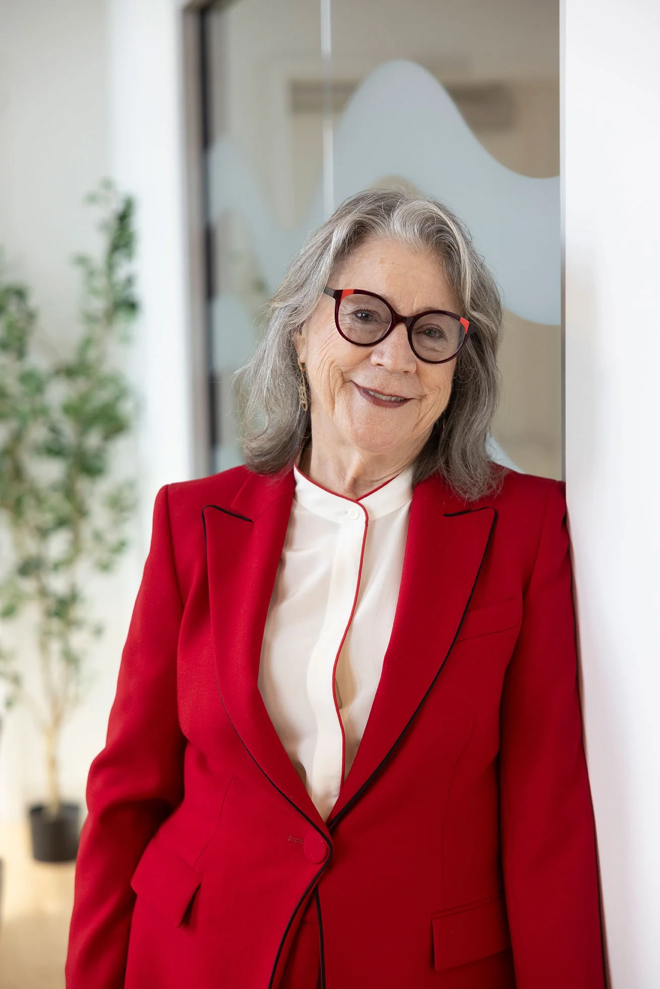 An older woman with gray hair and glasses, wearing a red blazer over a white blouse, smiling and leaning against a white wall.