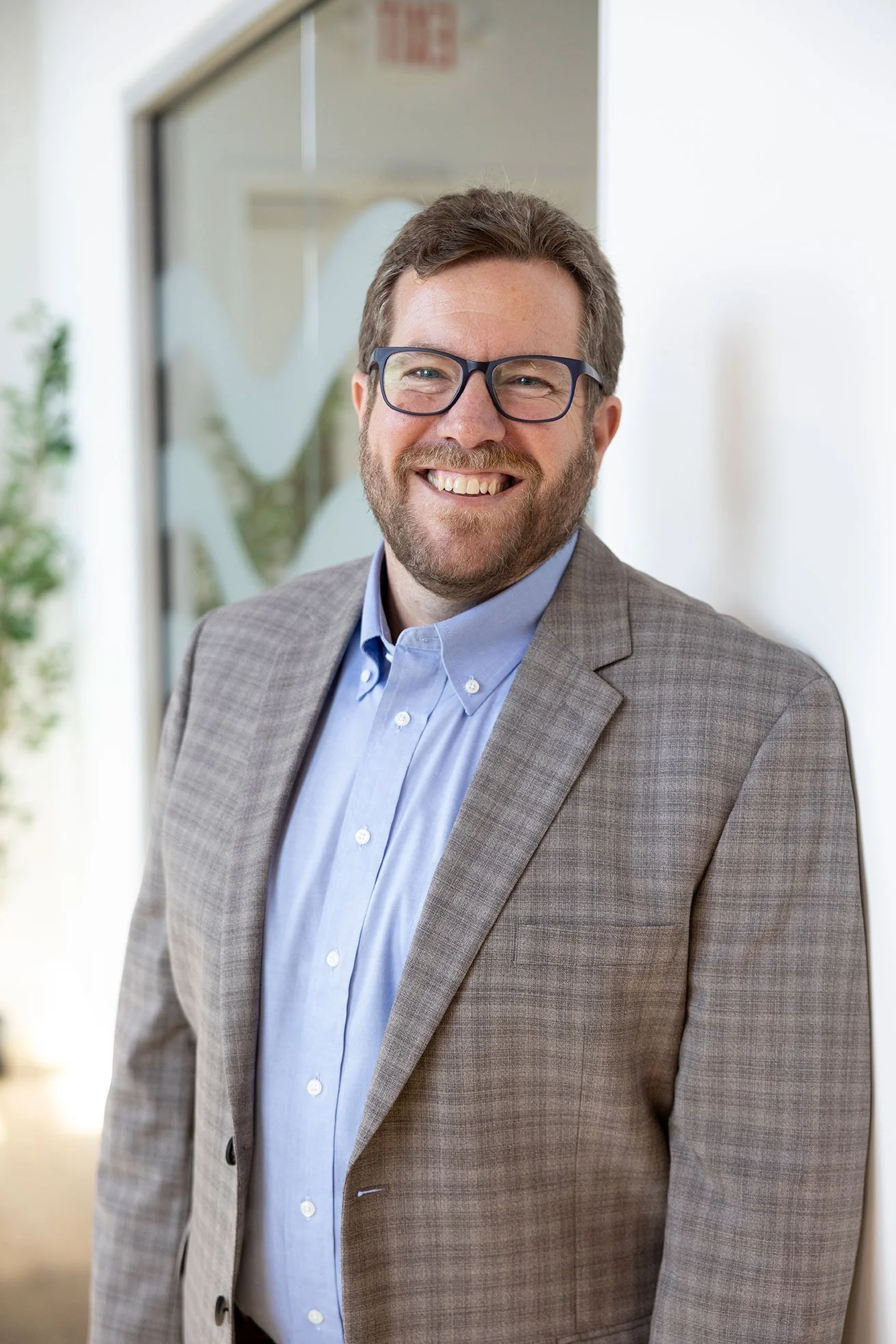 Portrait of a smiling man wearing glasses, a light blue button-up shirt, and a gray suit jacket, standing in an indoor setting with a glass window and a white wall in the background.