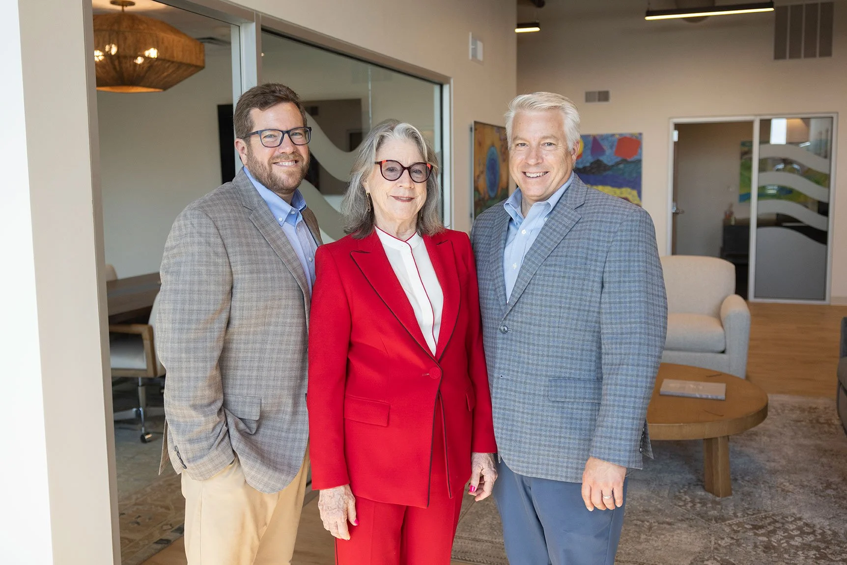 Three people, two men and one woman, standing together in a living room, smiling at the camera. The woman in the center is wearing a red suit and glasses. The man on the left has a beard and glasses, dressed in a checked blazer and beige pants. The man on the right has white hair and is wearing a grey checkered blazer with light blue shirt. The room has artwork on the wall and modern furniture.