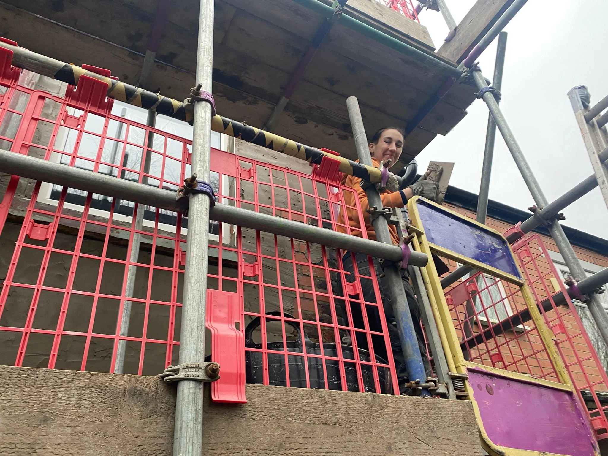 Naomi is on scaffolding while rendering the exterior of a house, holding a plastering trowel and smiling at the camera.