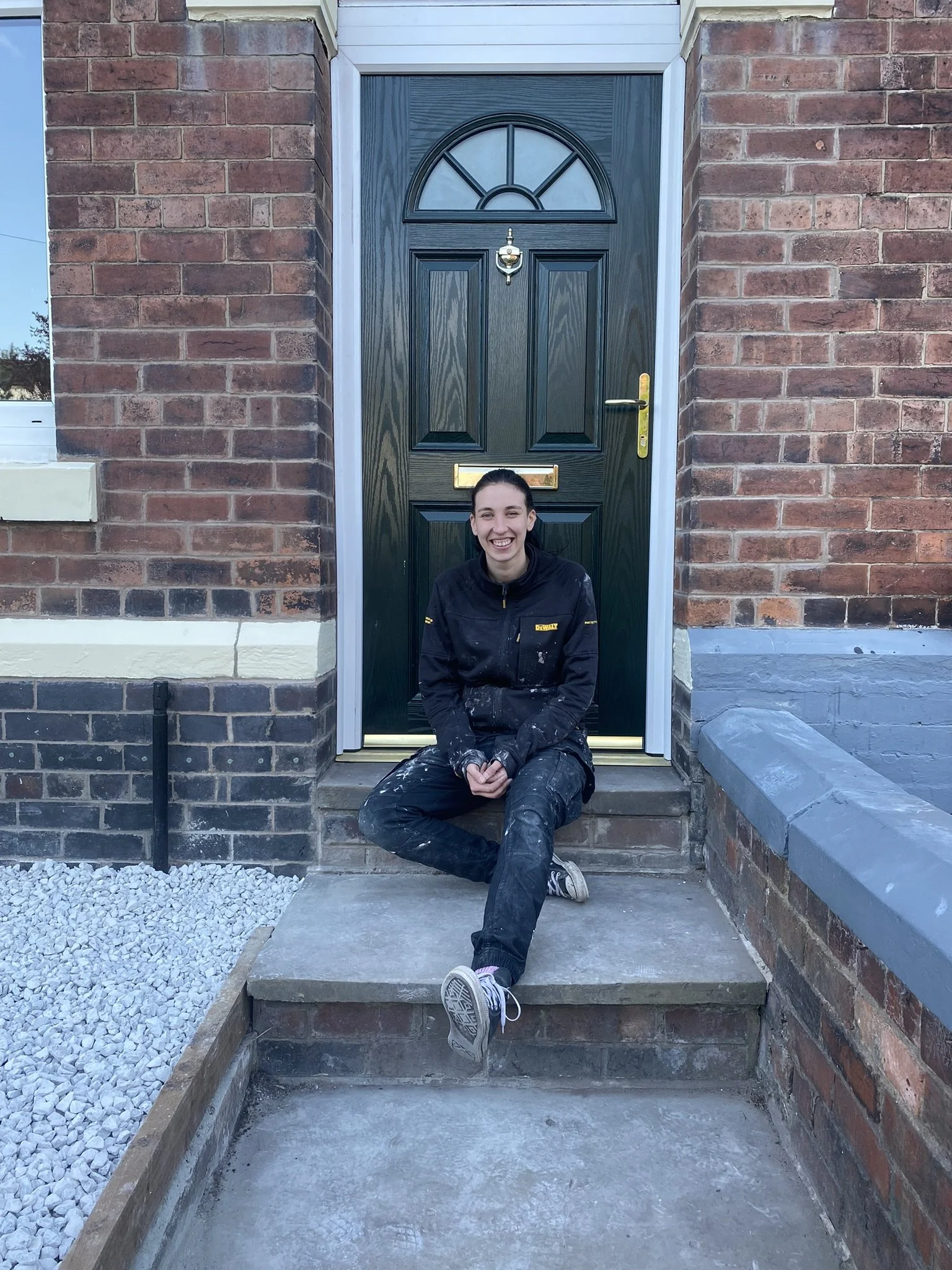 Naomi is sitting on the front step of a house, smiling at the camera. She has plaster on her work uniform