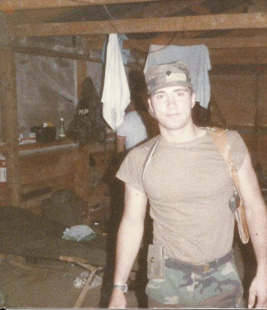 Steven Stinnett in his Army military camouflage pants, a tan t-shirt, wearing a camouflage cap, and a backpack, standing inside a wooden shelter during a deployment mission.