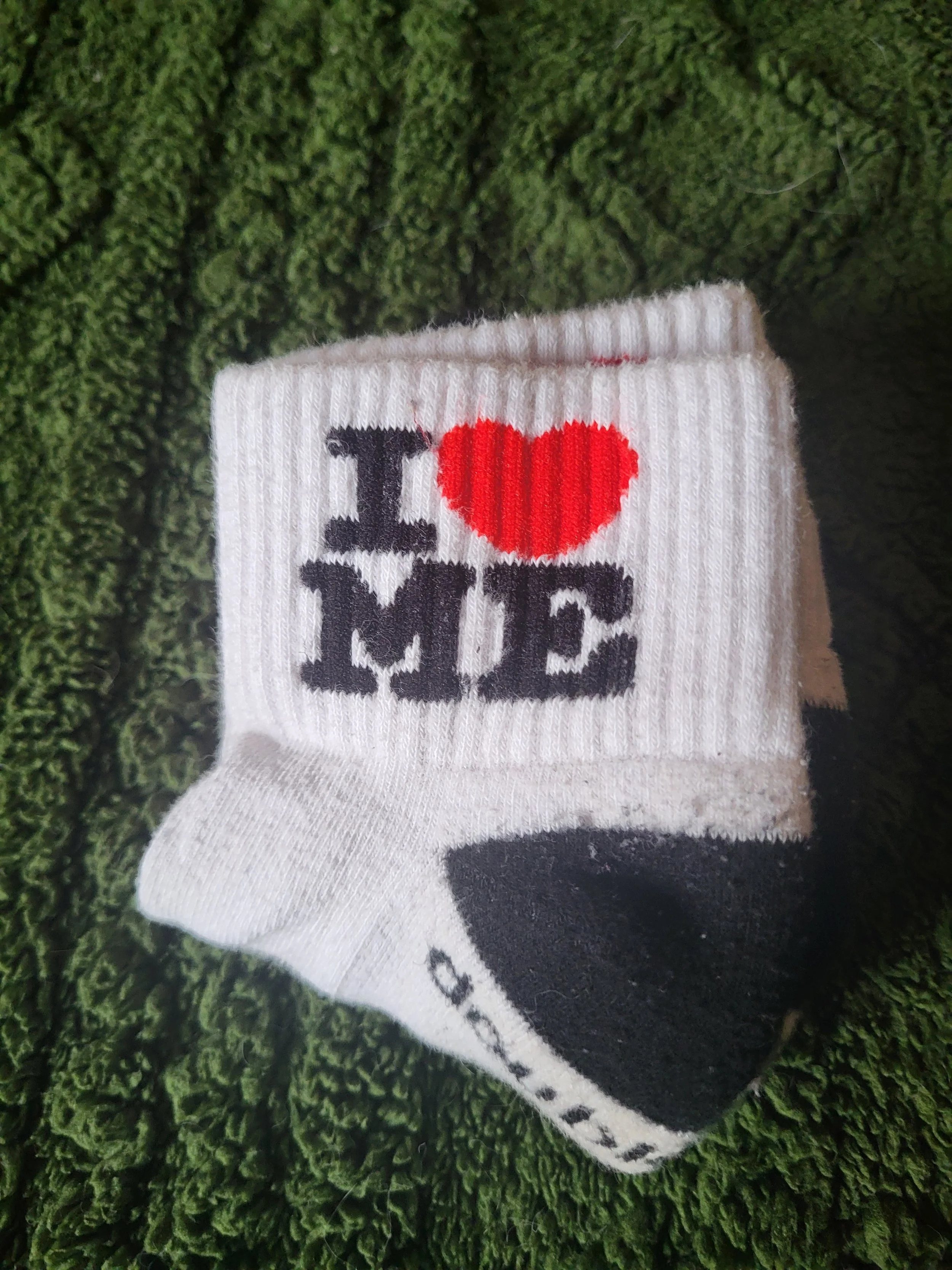 Close-up of a white sock with the word 'LOVE' and a red heart symbol on it, laid on dark green moss.