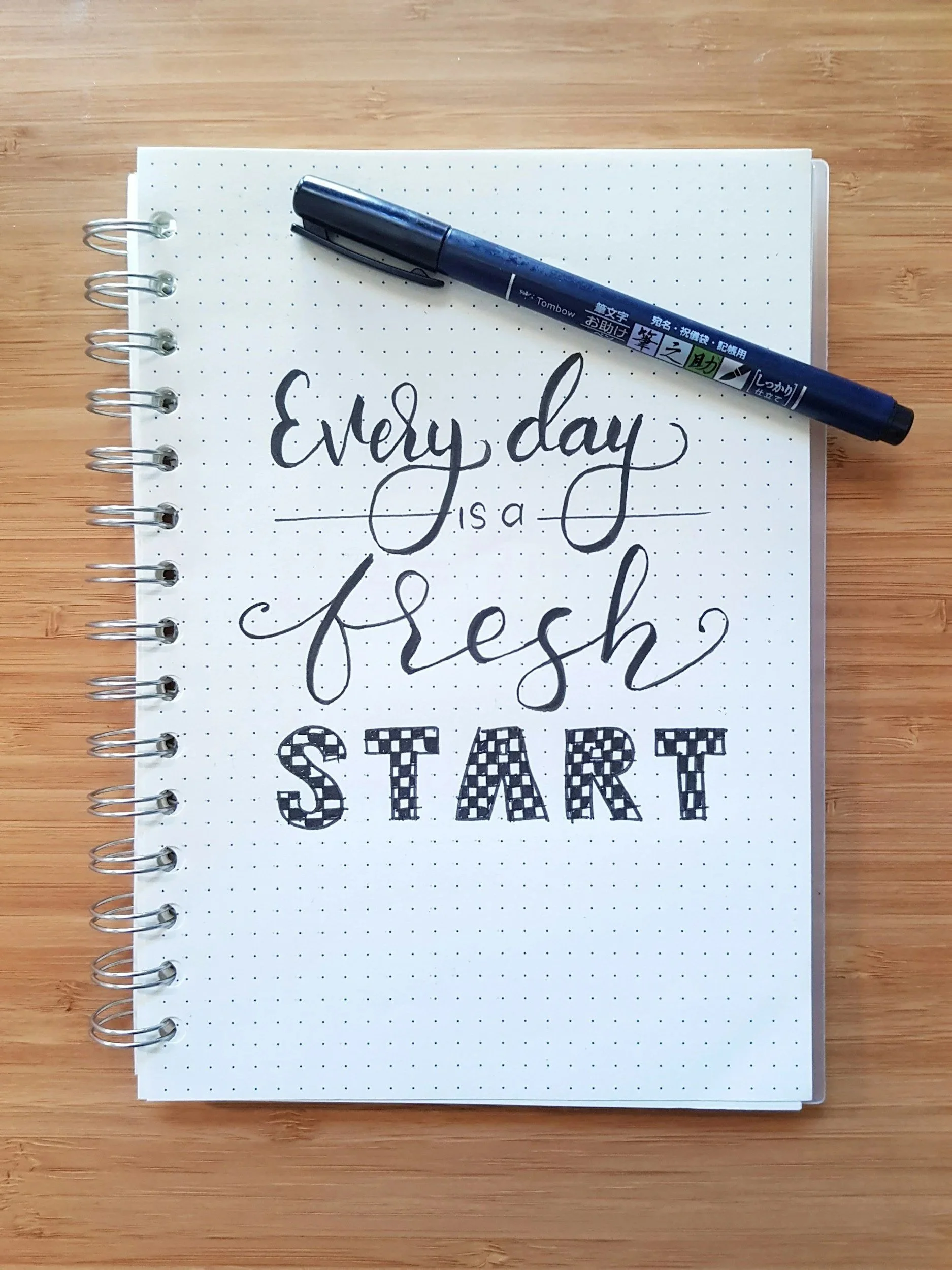 A spiral notebook with dotted pages on a wooden surface, featuring the handwritten message 'Every day is a fresh start' and a black marker resting on top.