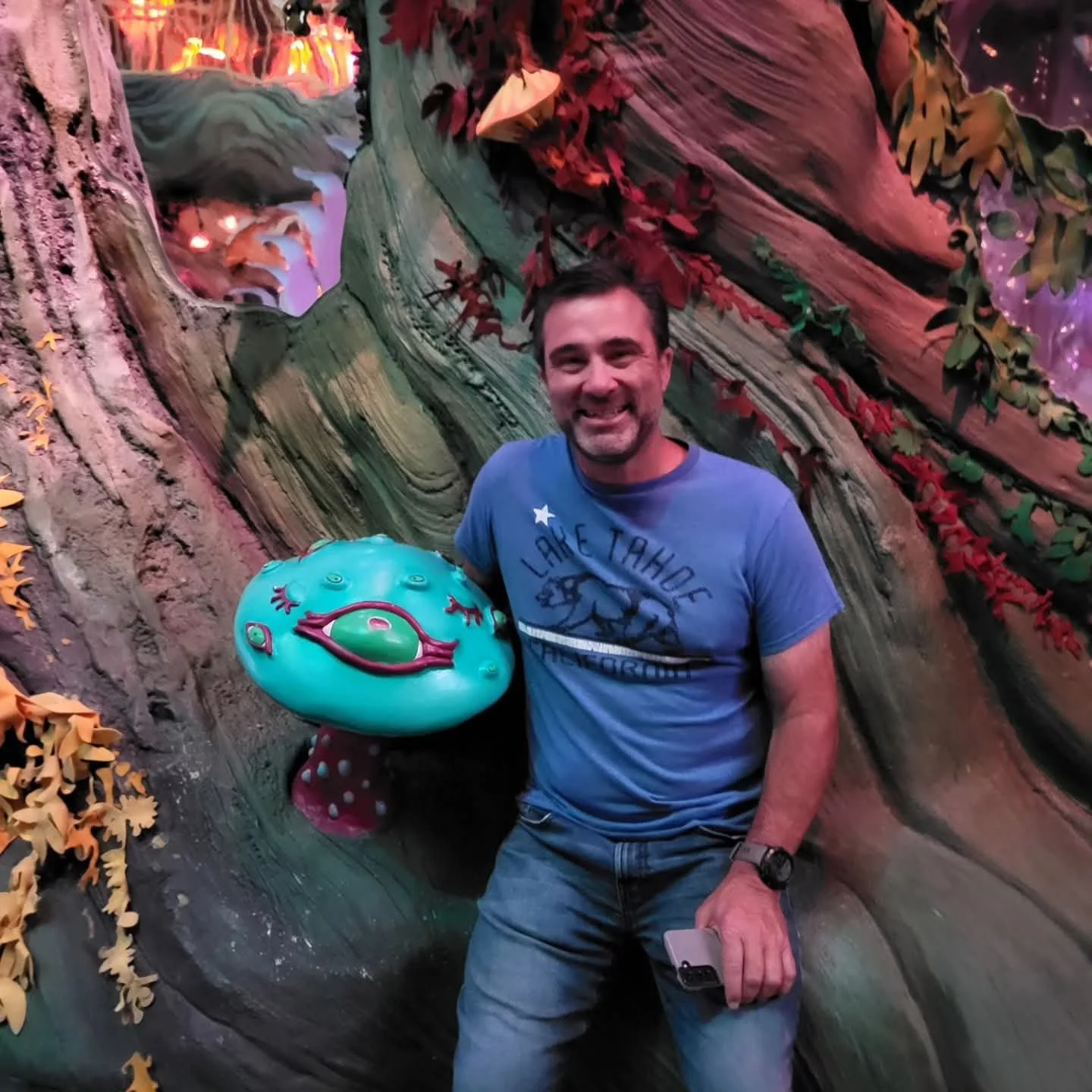 Steven Stinnett smiling while holding a colorful, cartoonish mushroom sculpture in front of a painted forest backdrop.