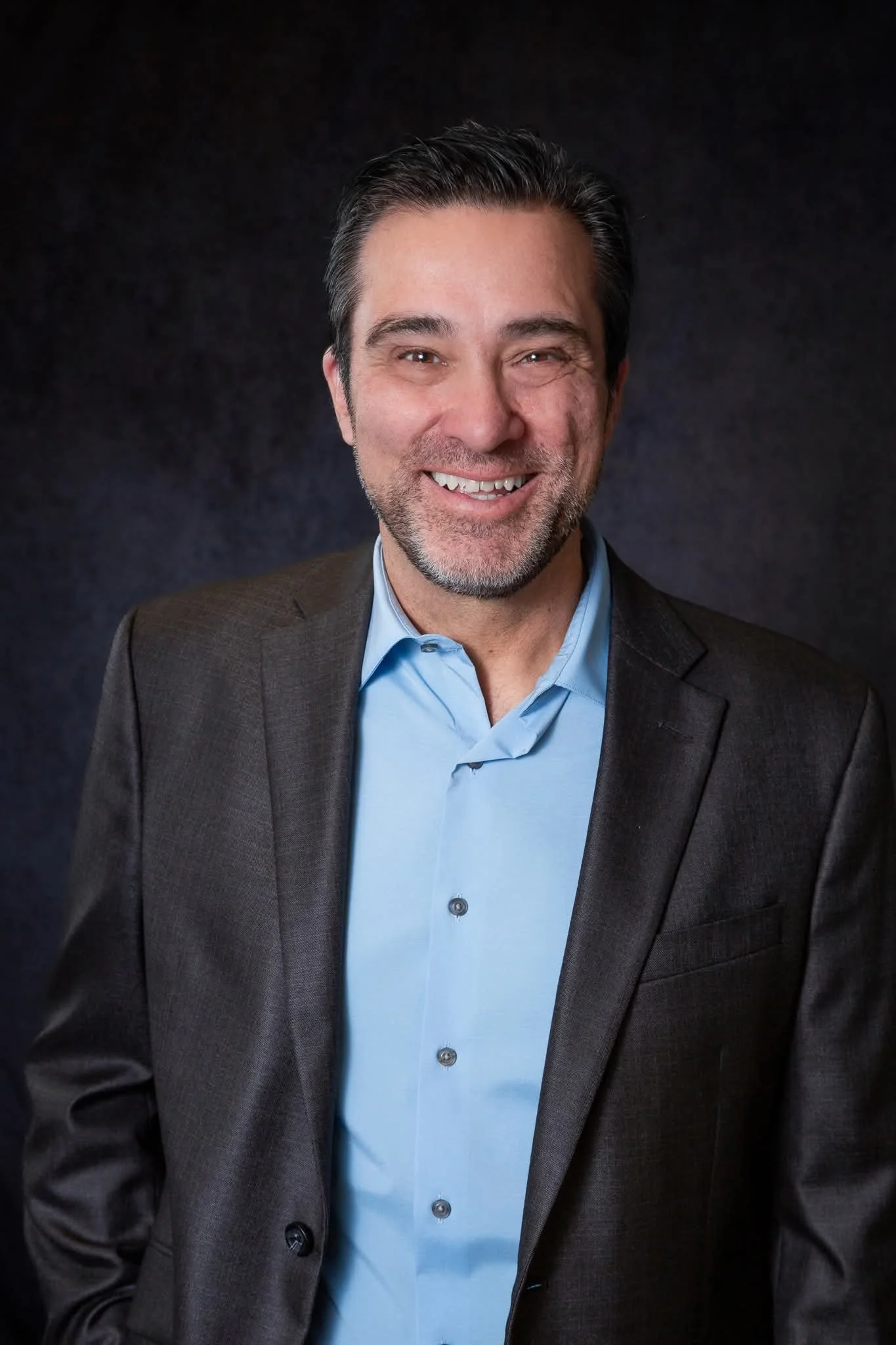 Steven Stinnett a middle-aged man with dark hair and a beard, wearing a dark suit and a light blue dress shirt, smiling against a plain dark background.