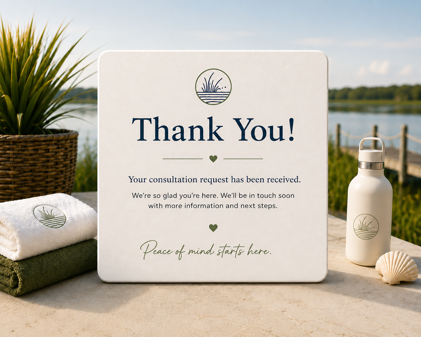A thank you sign on an outdoor table near a lake, with a white water bottle and neatly folded white and green towels, and a potted plant in the background.
