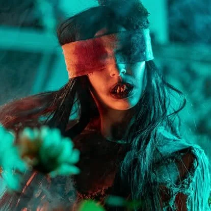 Blindfolded horror character surrounded by eerie flowers and atmospheric lighting, illustrating immersive storytelling, theatrical makeup, and psychological horror inside a haunted attraction.