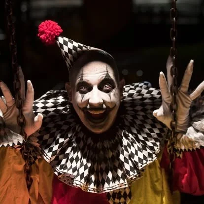 Sinister black-and-white harlequin clown performer emerging from darkness inside a haunted house attraction, showcasing detailed costume design, immersive entertainment, and Halloween horror experiences.
