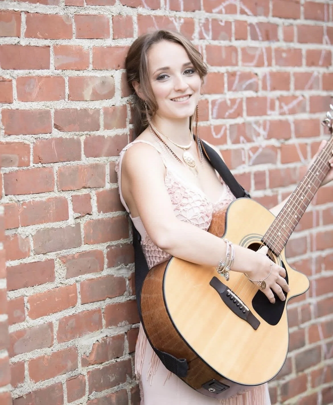 A young woman with brown hair and a pink dress stands against a red brick wall, smiling and playing an acoustic guitar.