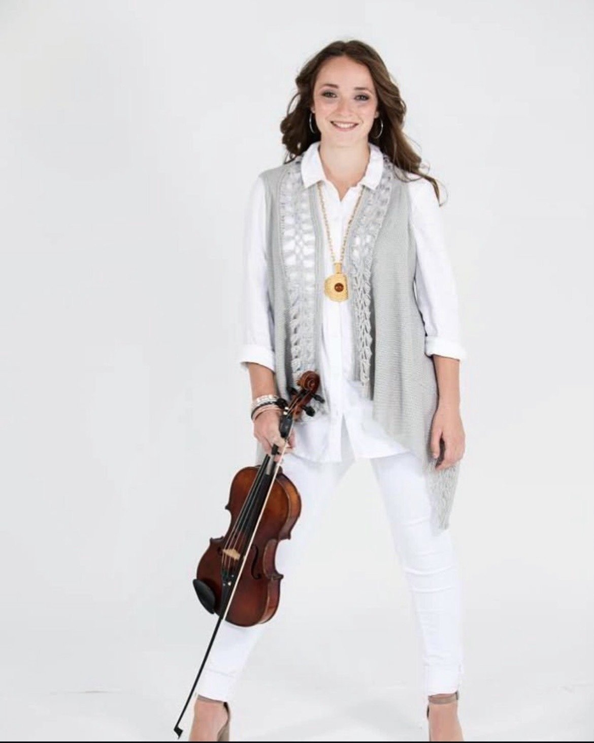 Woman dressed in white holding a violin, smiling against a plain white background.
