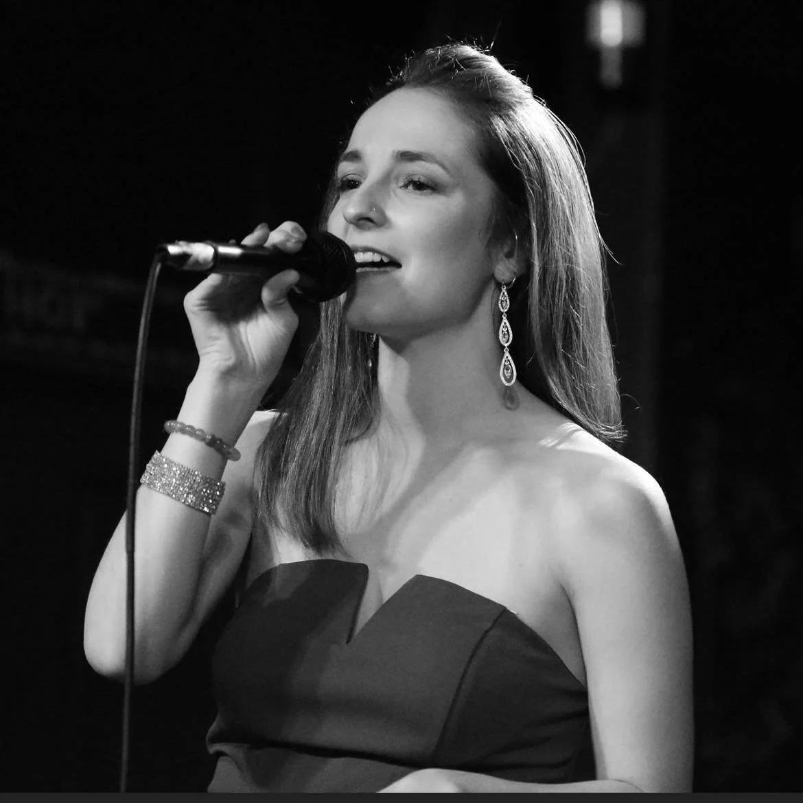 A young woman singing into a microphone on stage, wearing a strapless dress and long earrings, with a bracelet on her right wrist, in black and white.