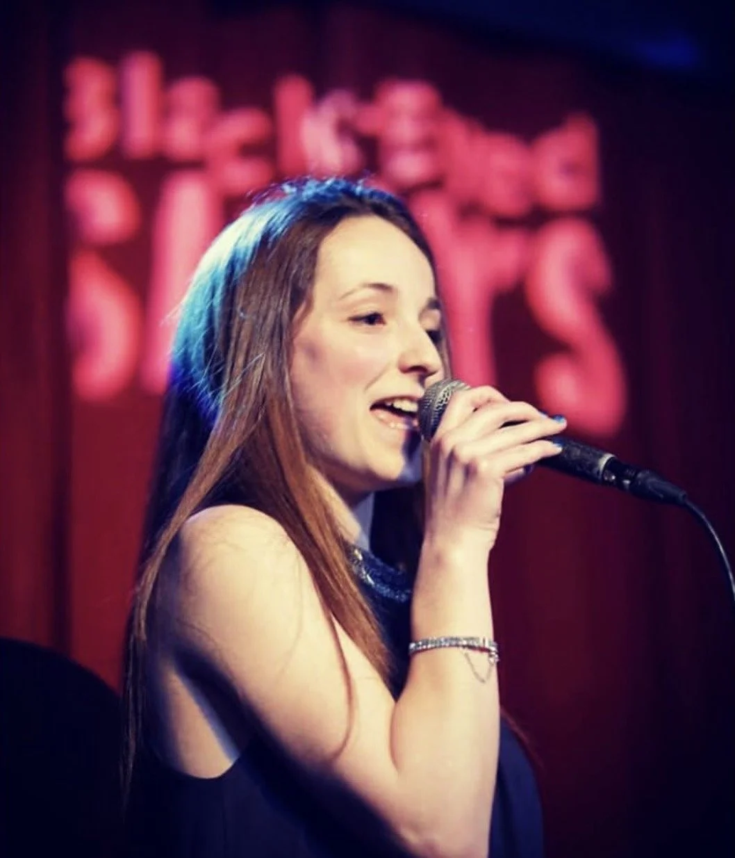 A woman with long reddish-brown hair singing into a microphone on stage, with a blurred pink and red background.