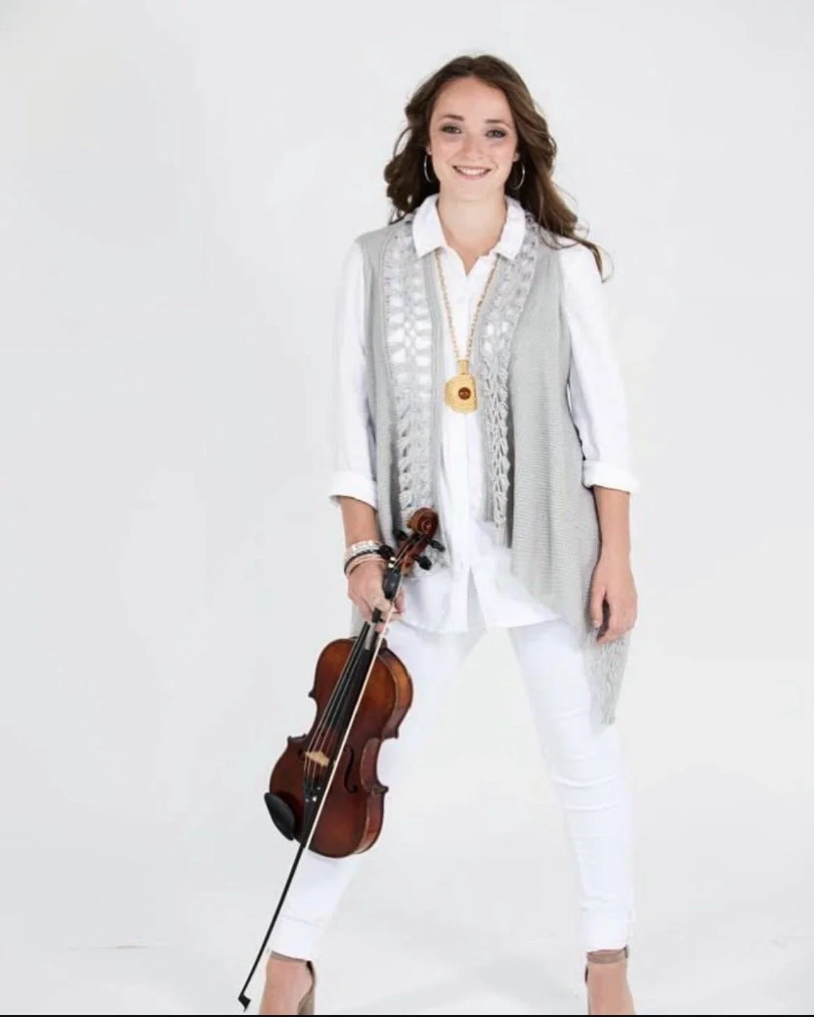 A woman with brown hair, wearing a white shirt, light gray vest, and white pants, holding a violin in her right hand, standing against a plain white background.