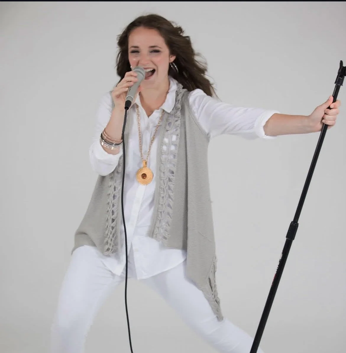 A woman singing into a microphone while holding a microphone stand, dressed in white, with a grey vest and accessorized with jewelry, standing against a plain background.
