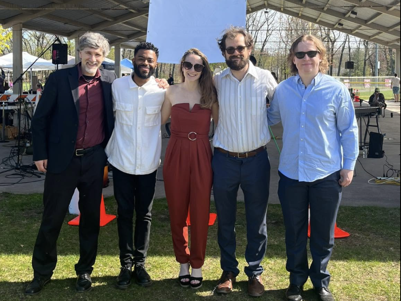 Five people standing together outdoors at an event, with a stage and musical equipment behind them. They are smiling and wearing casual to semi-formal clothing, with some wearing sunglasses.