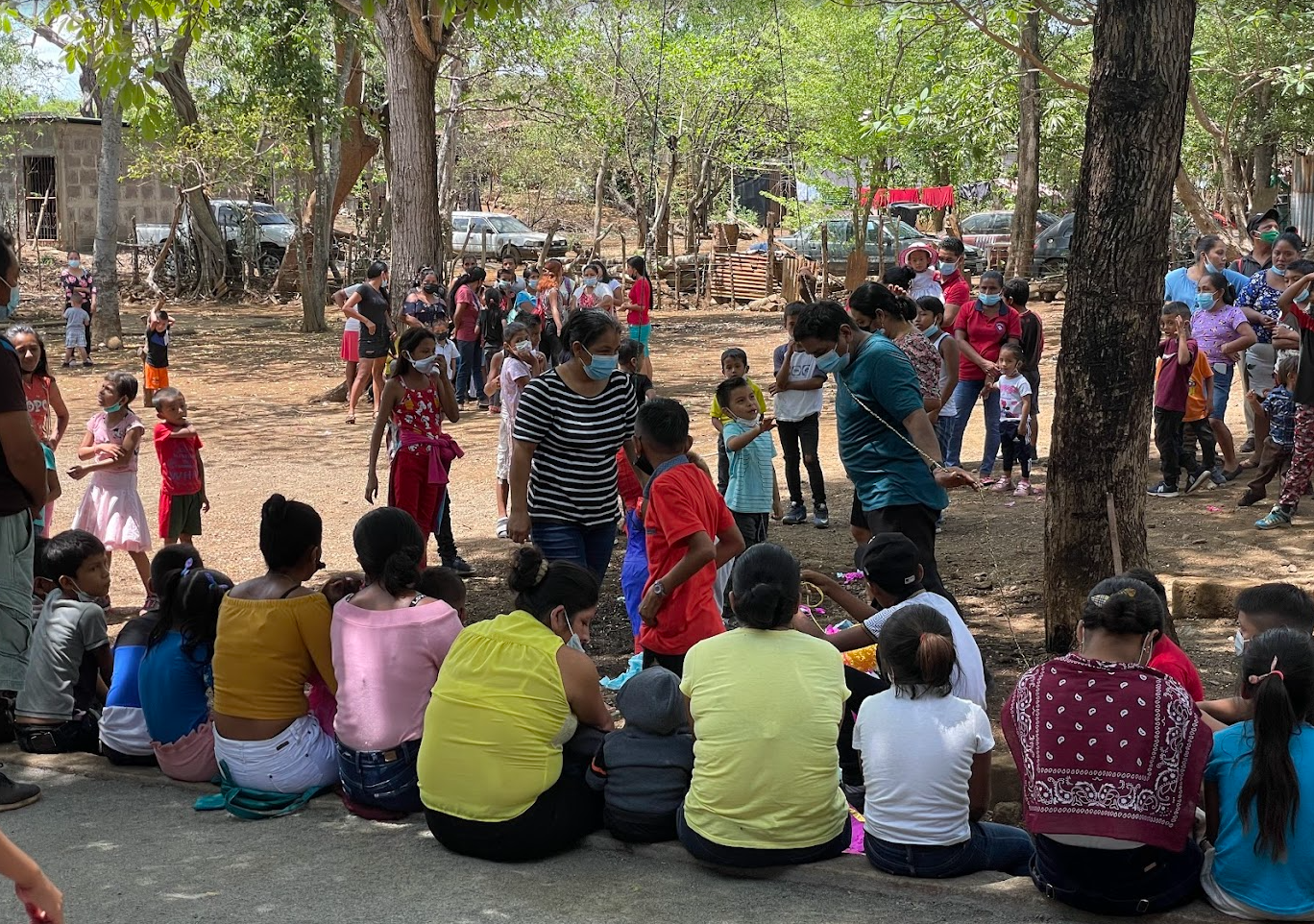 Group of adults and children gathered outdoors in a park under trees, some sitting and some standing, with many wearing face masks. The scene suggests a community event or gathering in a rural or semi-rural area.