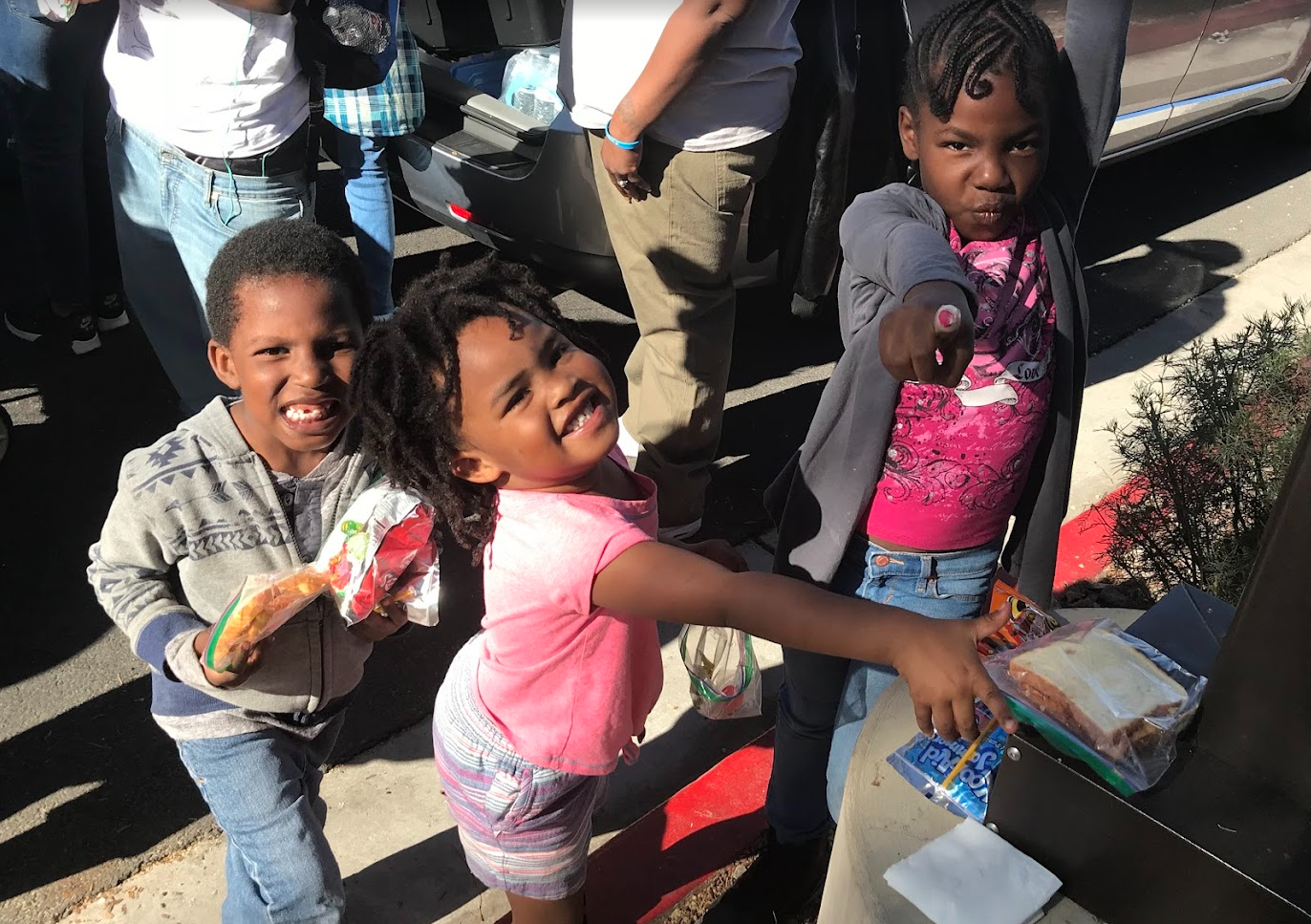 Three children standing in line outdoors, smiling and reaching out toward camera, with a woman partially visible behind them. The children appear happy and excited, with snacks visible in their hands.