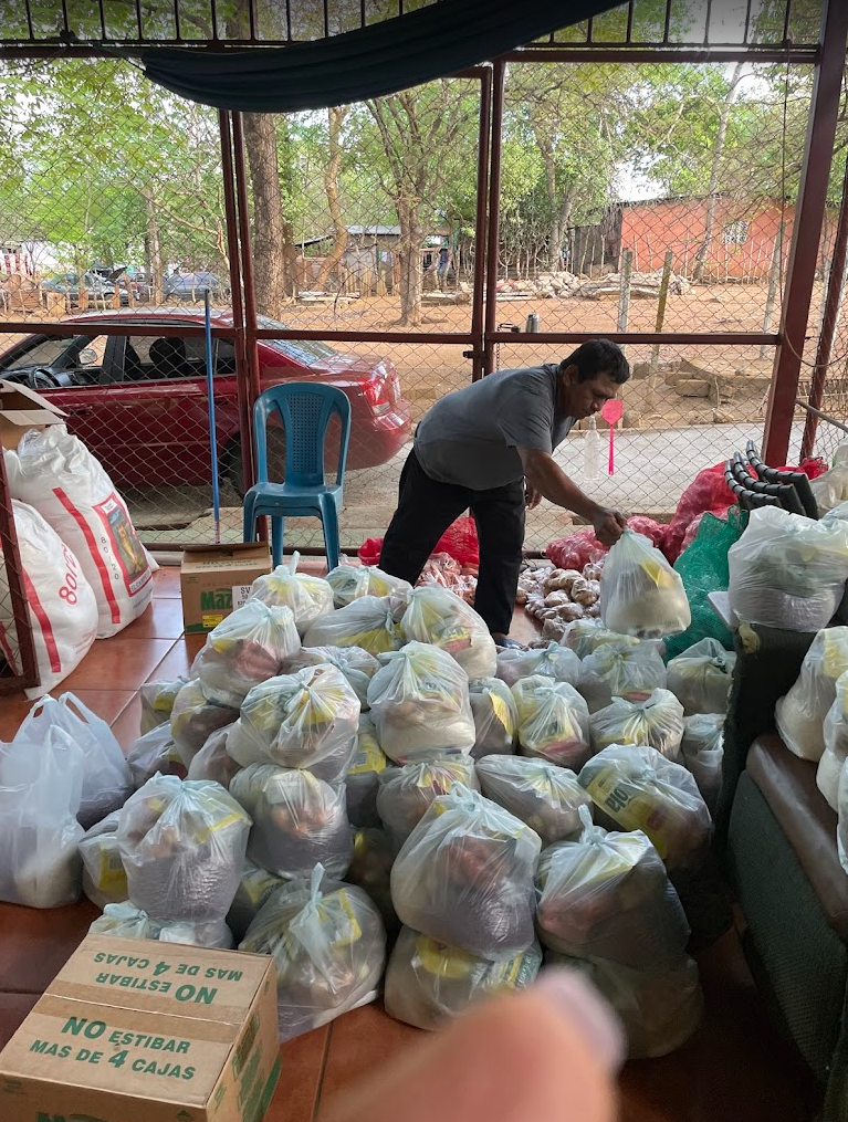 A man in a gray shirt and black pants organizes and bag groceries, with many plastic bags filled with groceries on the floor inside a covered outdoor area, with a chain-link fence, a red car, and trees outside in the background.
