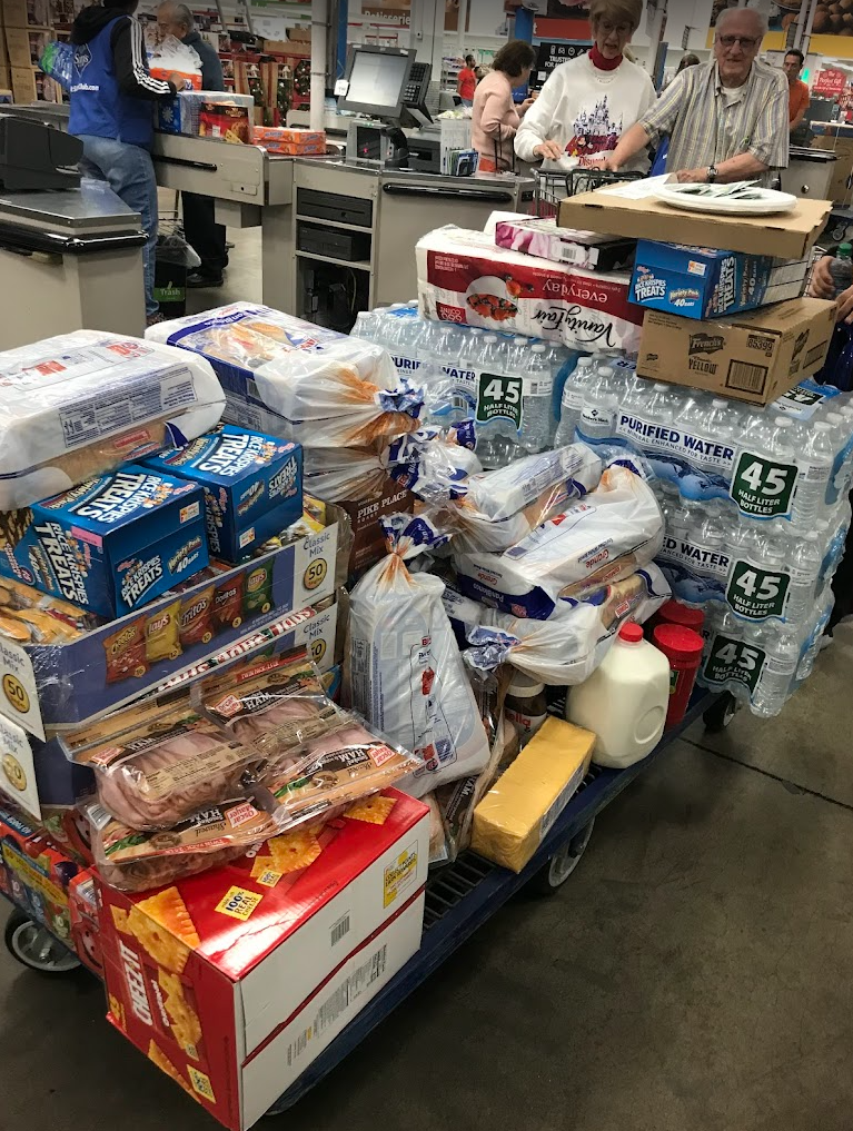A shopping cart in a grocery store filled with bottled water, bread, snacks, and other groceries, with checkout lanes and customers in the background.