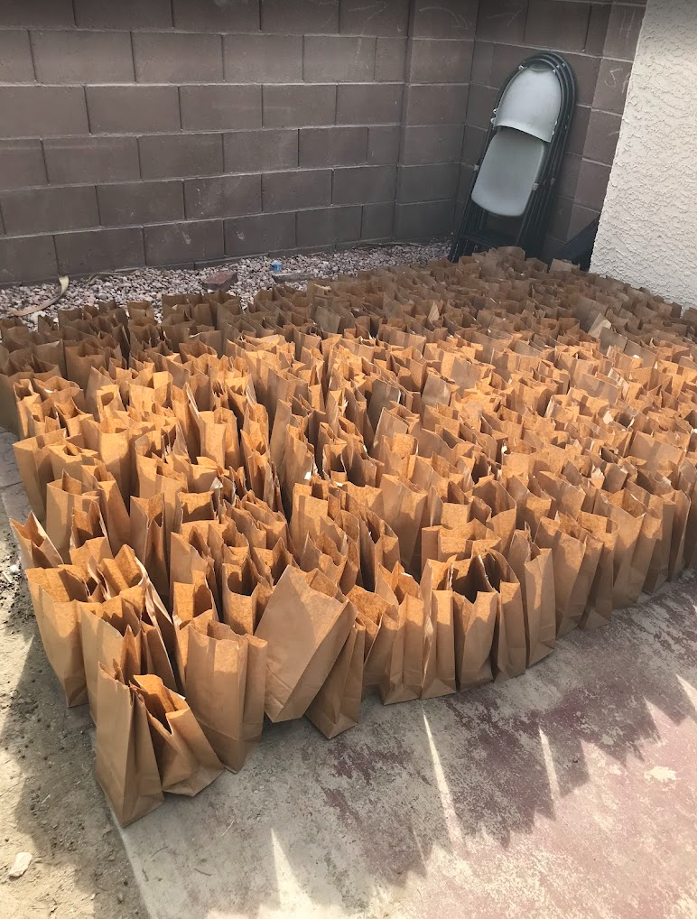 Numerous paper bags filled with items, arranged outside near a brick wall. Some metal chairs stacked in the corner.