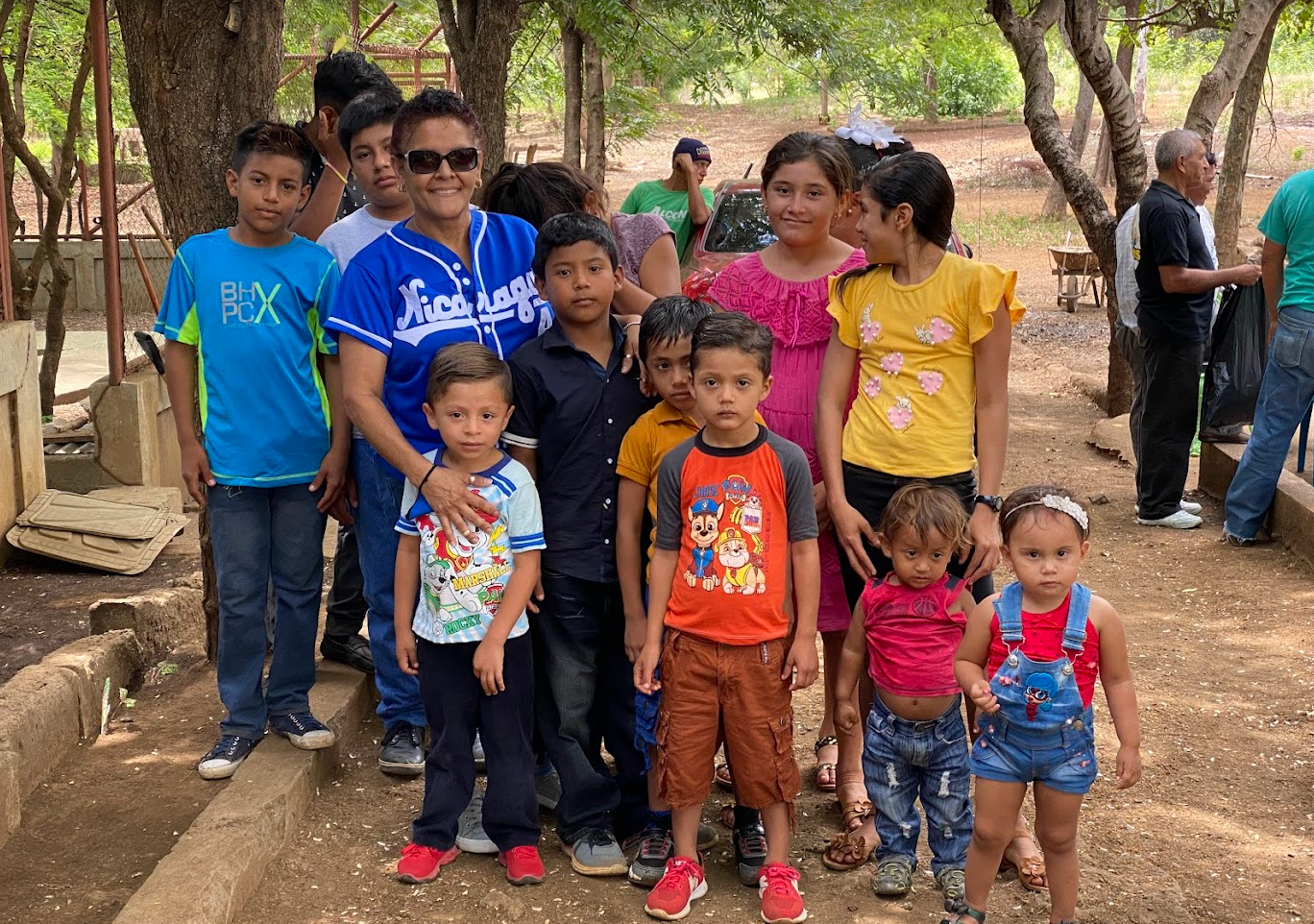A group of children and a woman posing outdoors under trees, with some individuals in the background.