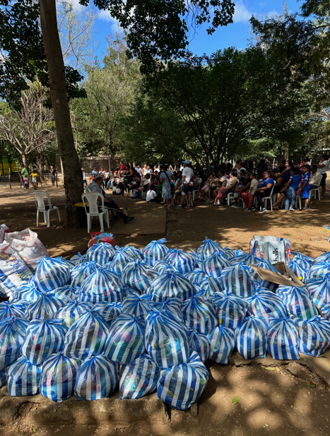 Numerous bags filled with supplies in the foreground, and a crowd of people seated on chairs and standing in a park area with trees in the background.