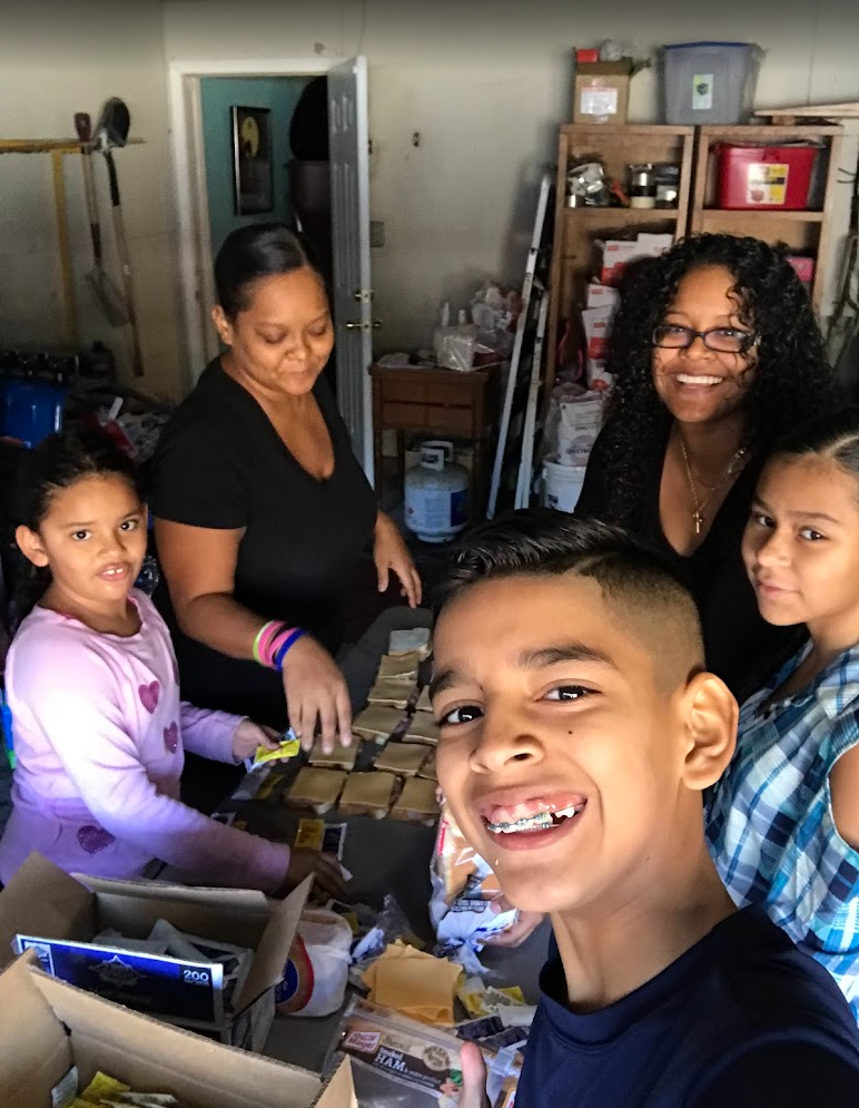 Five smiling people, including a young boy in the foreground, a girl in a plaid shirt, an older woman, and two women, around a table with food packaging in a garage or storage-room with shelves and tools in the background.
