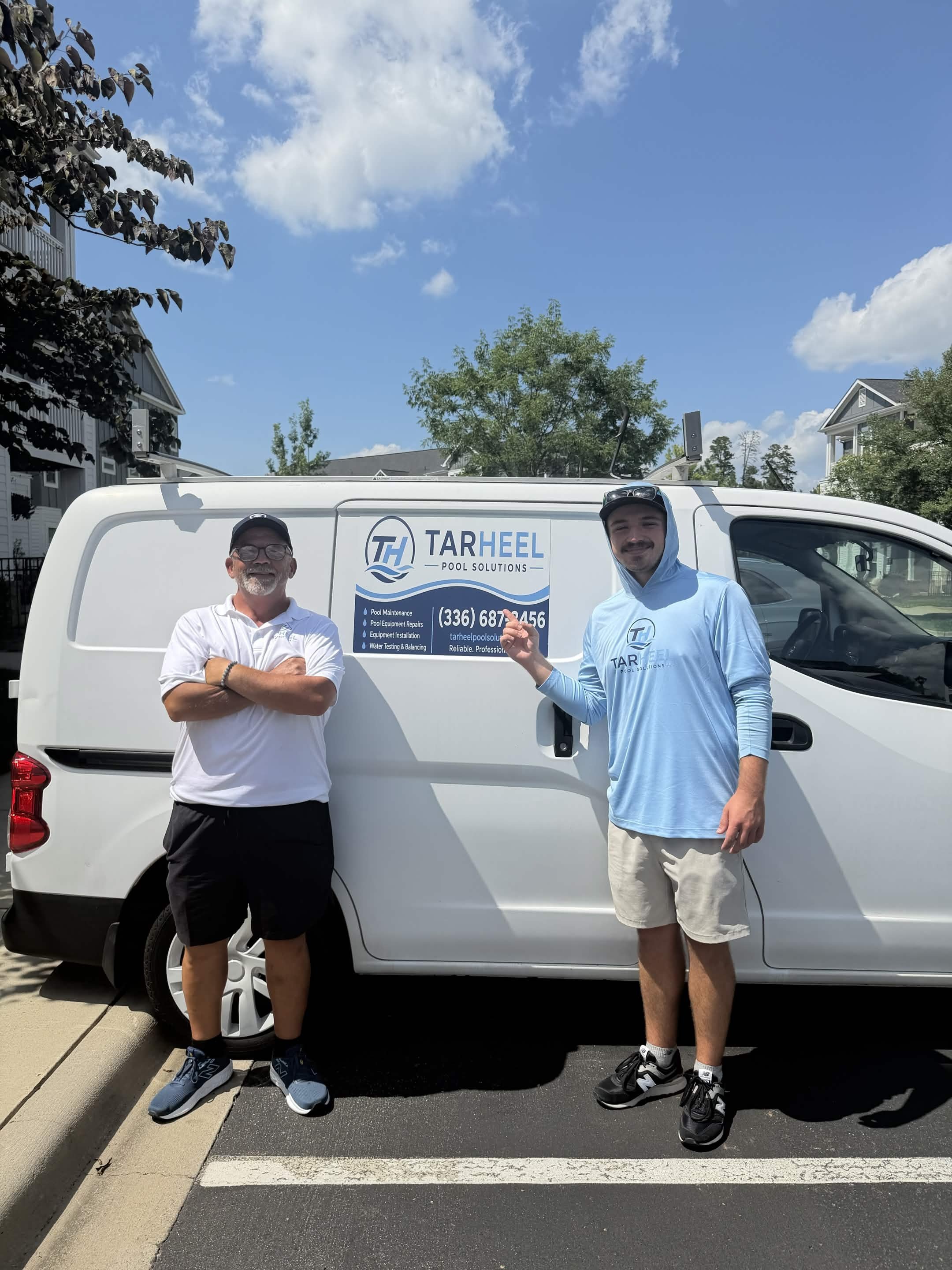 Two men standing in front of a white van with a company logo and phone number. One man is wearing a white polo shirt and black shorts, the other is wearing a light blue hoodie and beige shorts. The sky is blue with a few clouds, and there are houses and trees in the background.