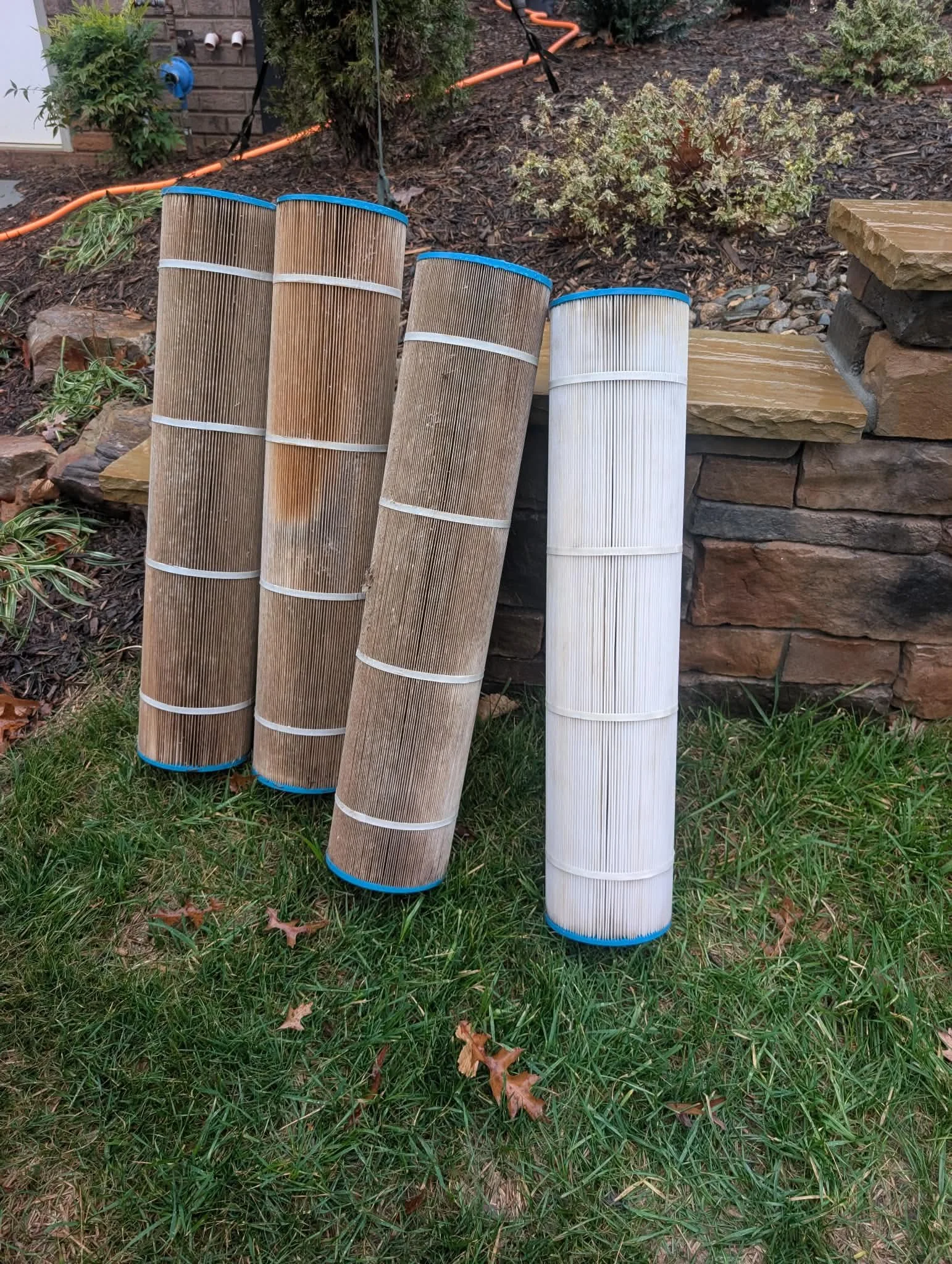 Four pool filters leaning against a stone wall and grass, with bushes and an orange garden hose in the background.