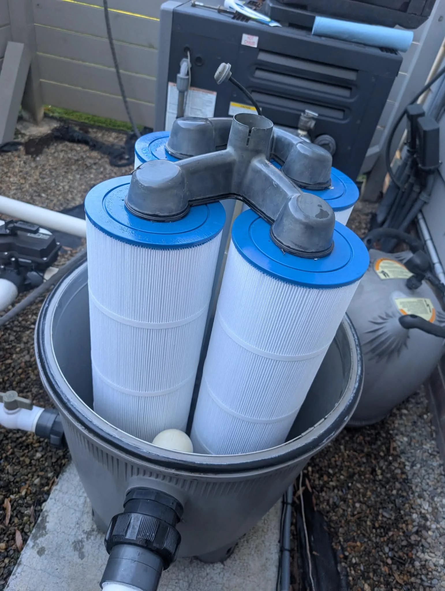 Pool filtration system with three large cylindrical filters connected by black pipes, located outdoors on a concrete surface near a grey fence and a black control box.