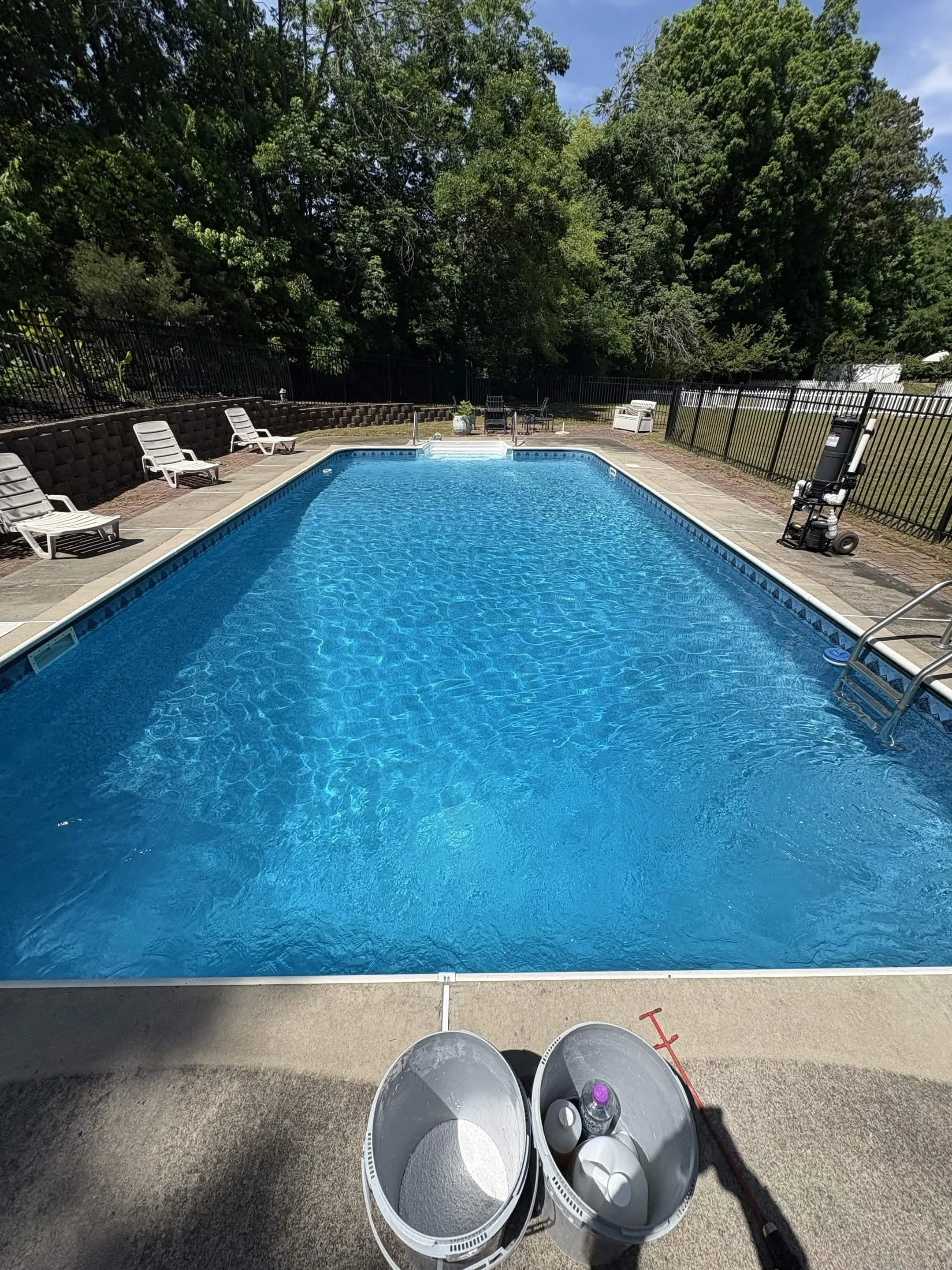 View of a backyard swimming pool with cleaning equipment, buckets, and seating area surrounded by a black fence and green trees.