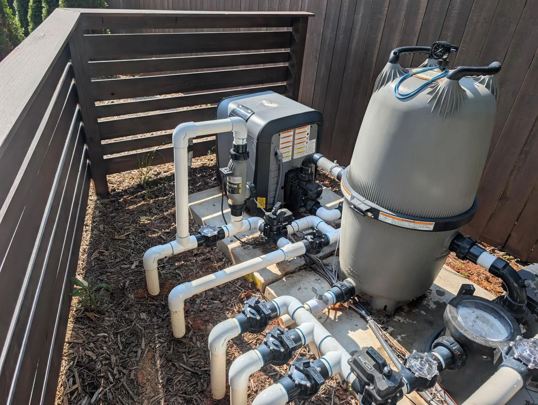An outdoor pool filter system with white pipes, a large gray filter tank, and a control box, installed on a concrete slab next to a wooden privacy fence, with mulch and small plants nearby.
