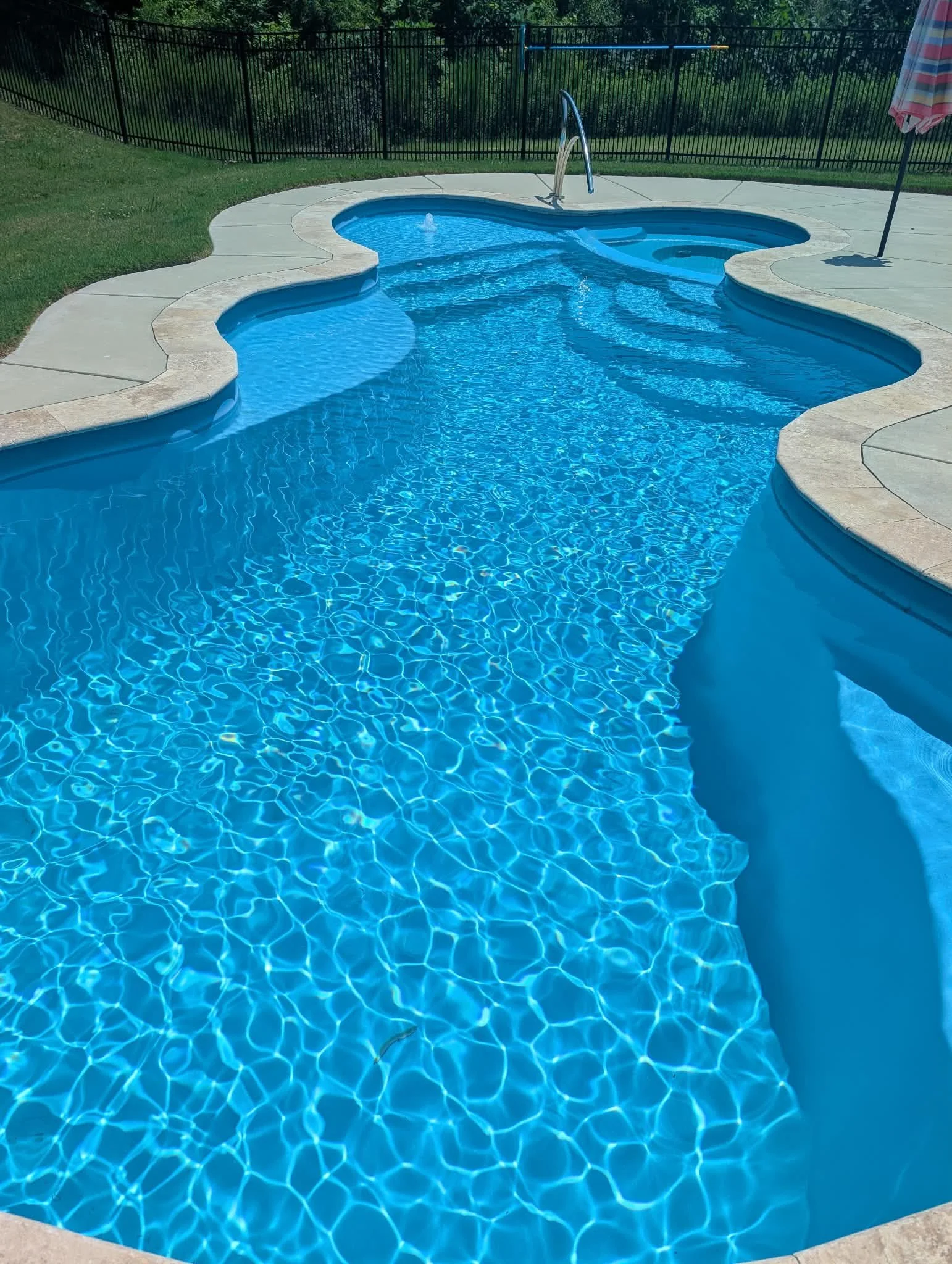 A backyard swimming pool with a kidney shape, surrounded by a concrete deck and black metal fence, with a striped umbrella on the right side.