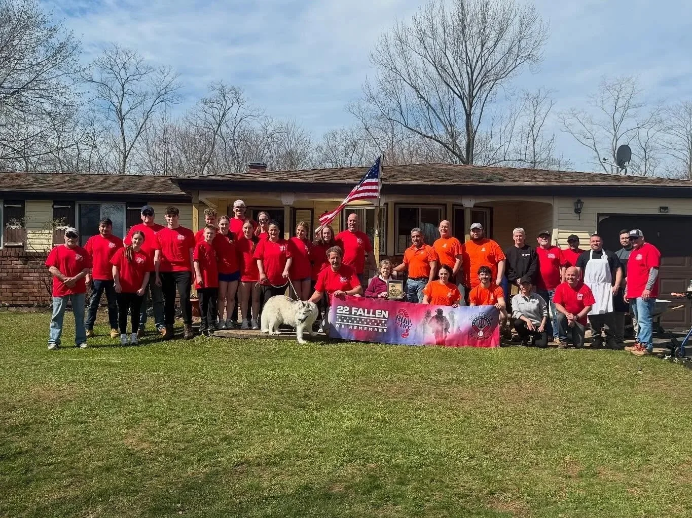 HNR Restorations volunteers for Act of Valor. large group of people gather in front of a house, with some holding a banner that says '22 Fallen We Remember'. They wear red and orange shirts, with a dog on a leash. An